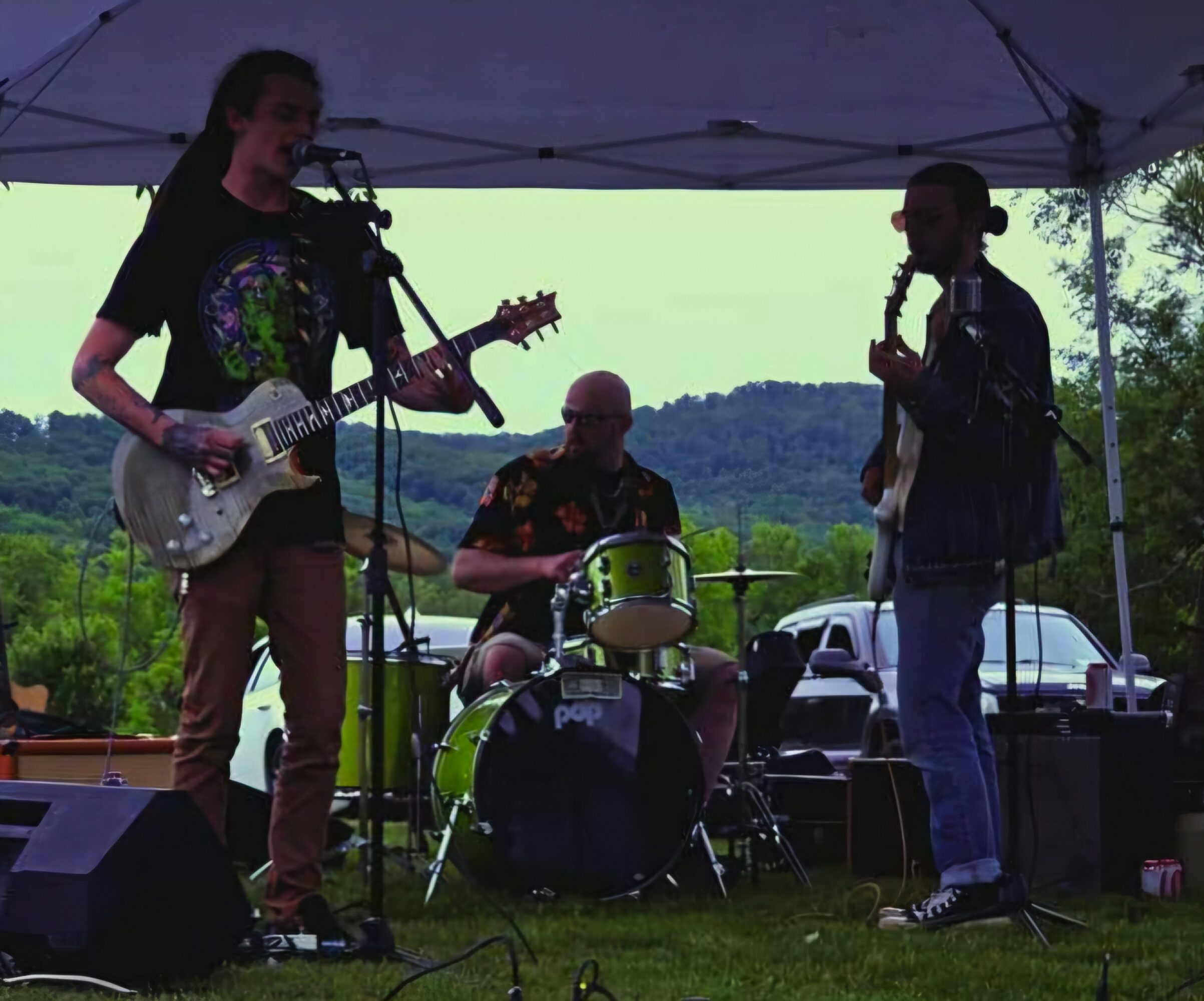 Three musicians perform outdoors under a canopy, with trees and hills in the background, playing guitar, drums, and singing.