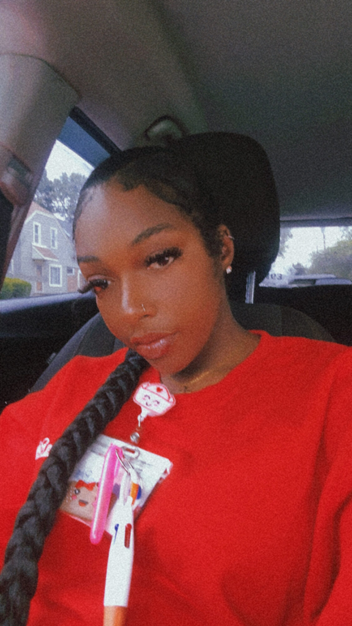 Young woman with long braided hair in a red shirt sitting in a car, looking at the camera.