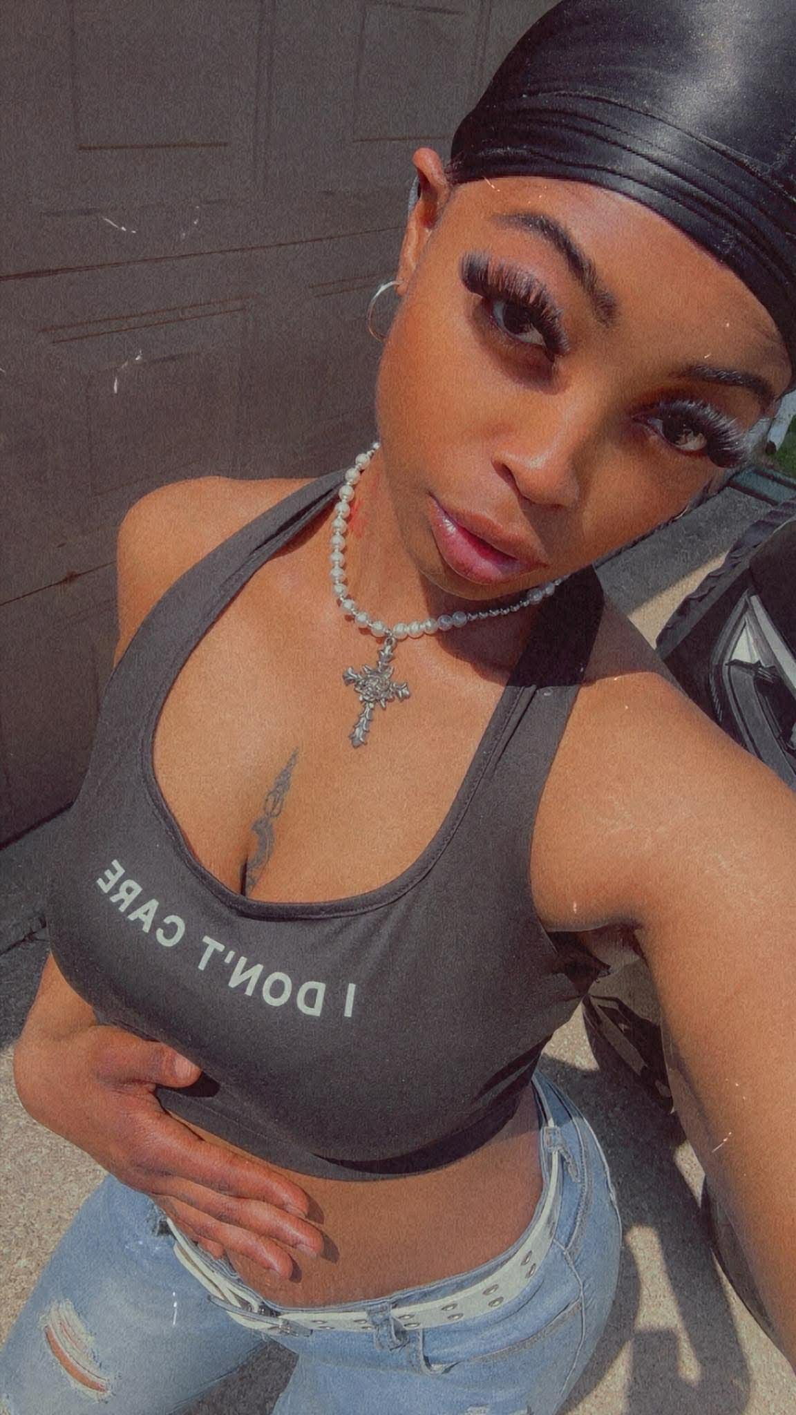 Woman with a headscarf, wearing a tank top and necklace, taking a selfie outdoors.