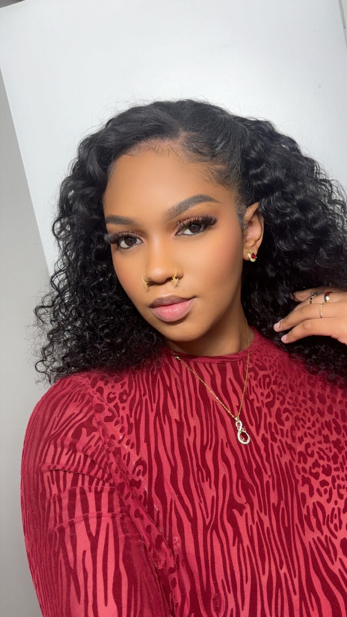 Young woman with curly hair wearing a red patterned top, looking at the camera, touching her hair, neutral background.