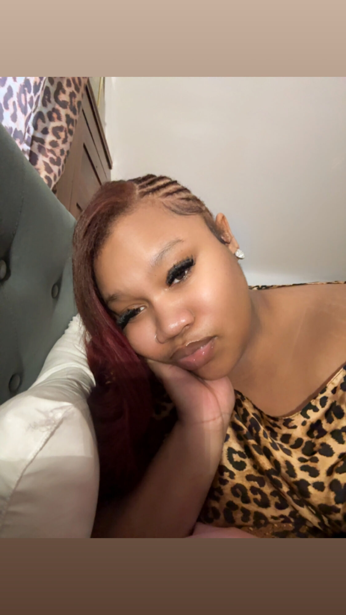 Young woman with long, reddish-brown hair lying on a bed, wearing a leopard print top, resting her face on her hand.