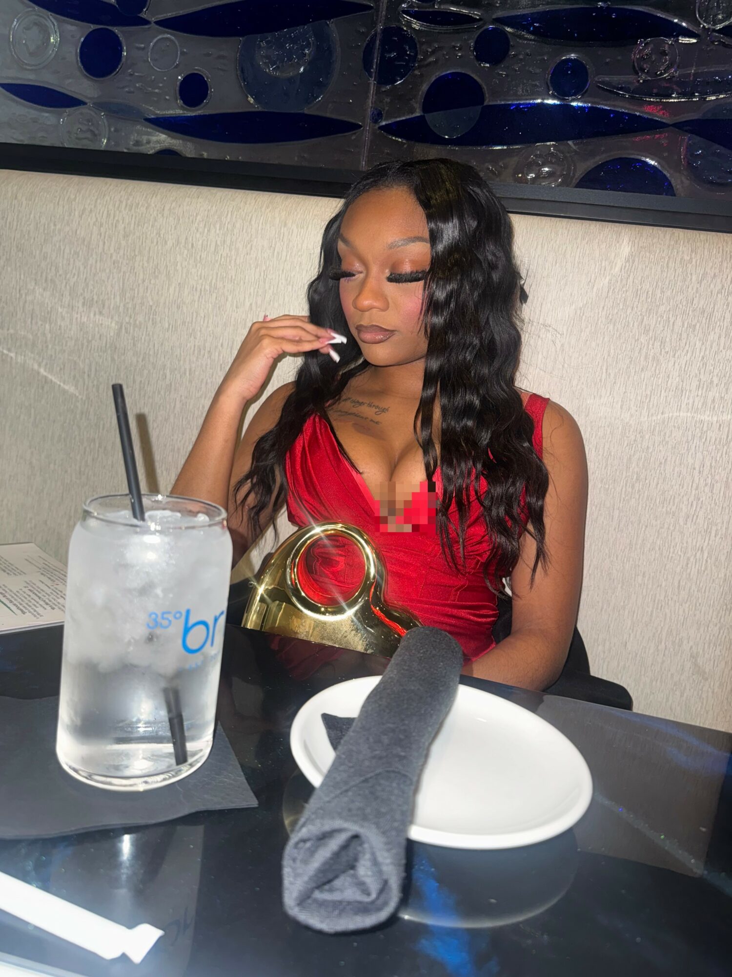 Woman in red dress sitting at a table with a glass of water, a napkin, and a plate, in a restaurant.