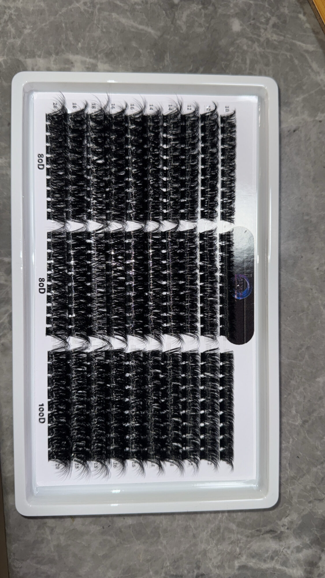Tray of black hair extensions or wigs arranged in rows inside a plastic container on a gray surface.