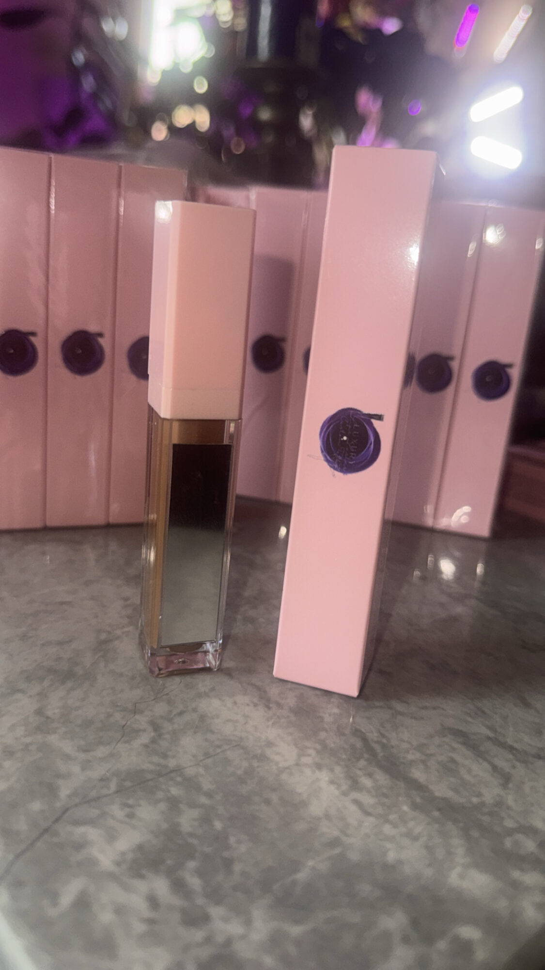 A clear glass perfume bottle with a pink box beside it, both on a gray surface, with blurred colorful lights in background.