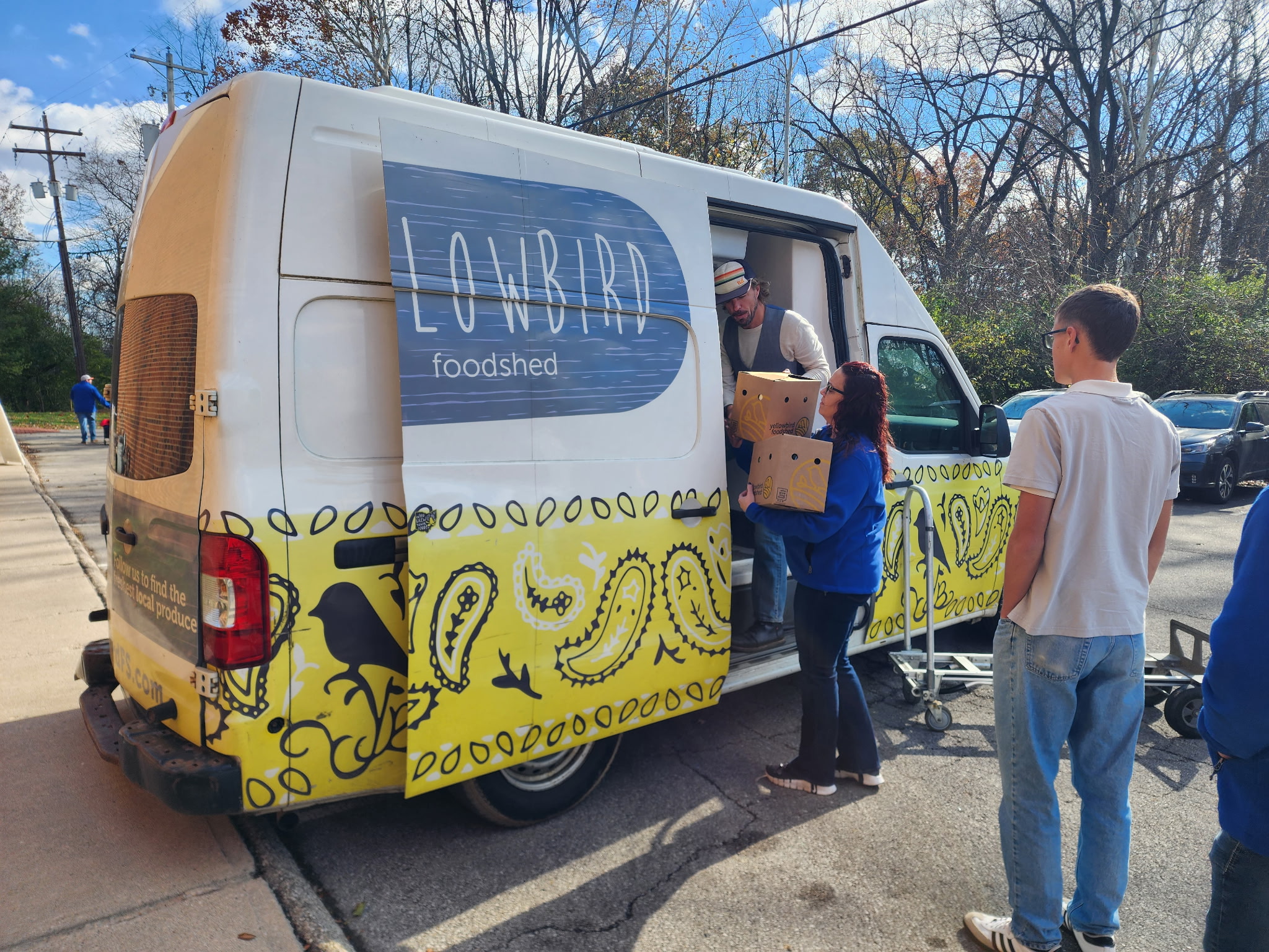 People line up at a food truck with a yellow and black design, and a sign reading 'LOUVIRD foodshed' on the side.