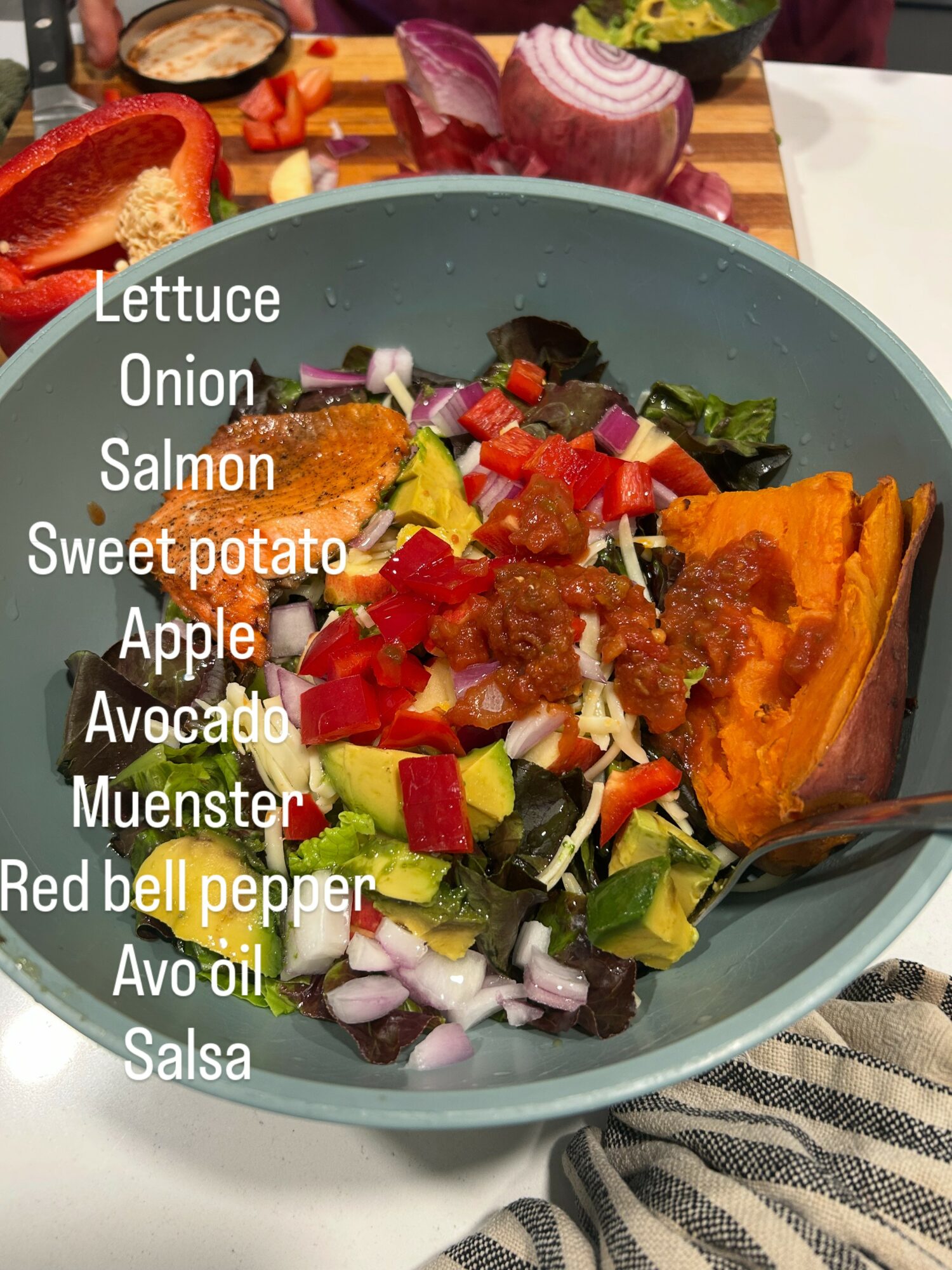 Bowl of salad with lettuce, onion, salmon, sweet potato, apple, avocado, red bell pepper, and salsa, with a spoon inside.