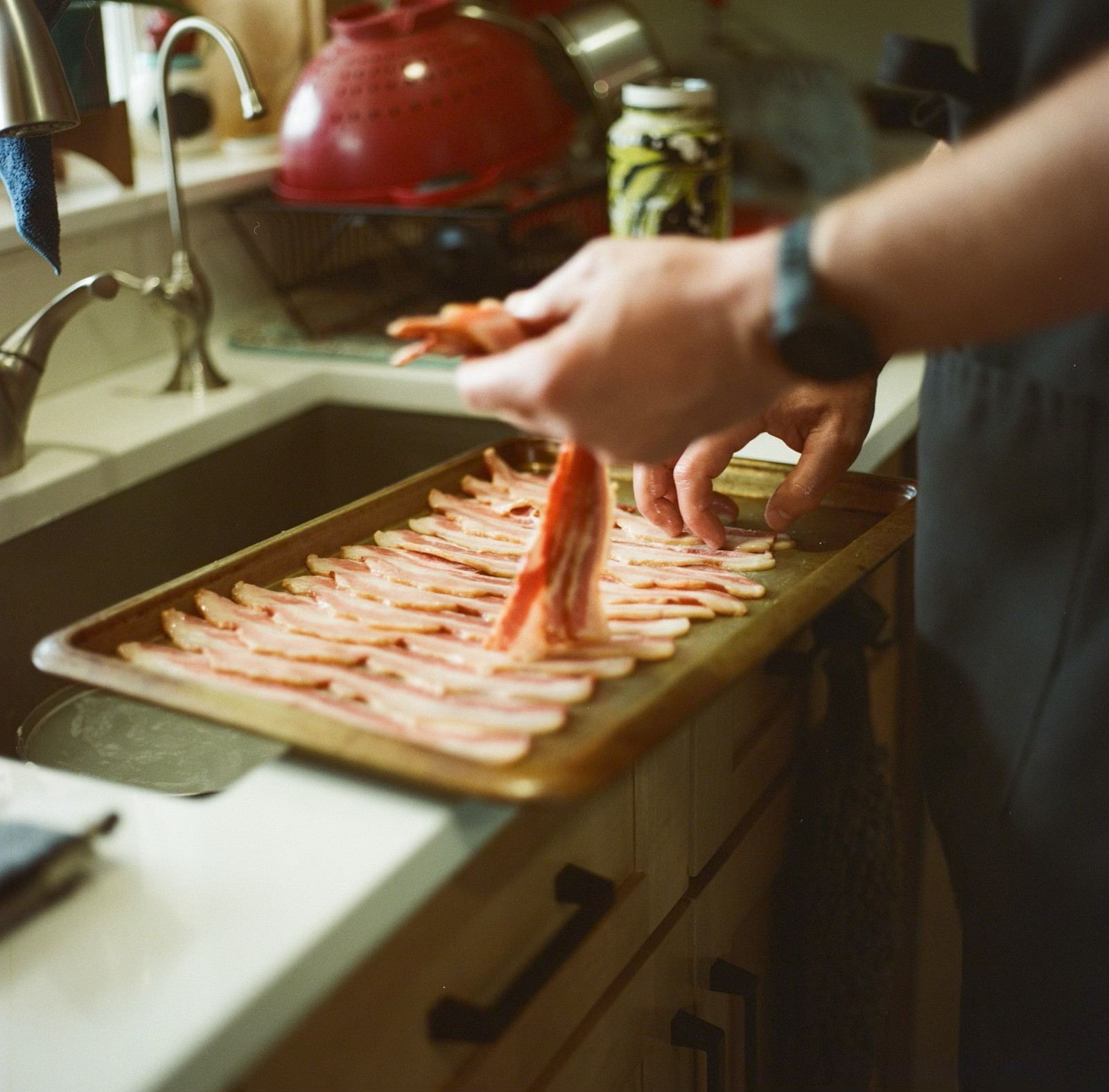 Person preparing bacon strips on a wooden cutting board in a kitchen, with a sink and red kettle in background.