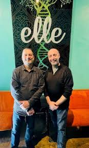 Two people stand in front of a backdrop with the word 'elle' and a DNA strand, smiling at the camera.
