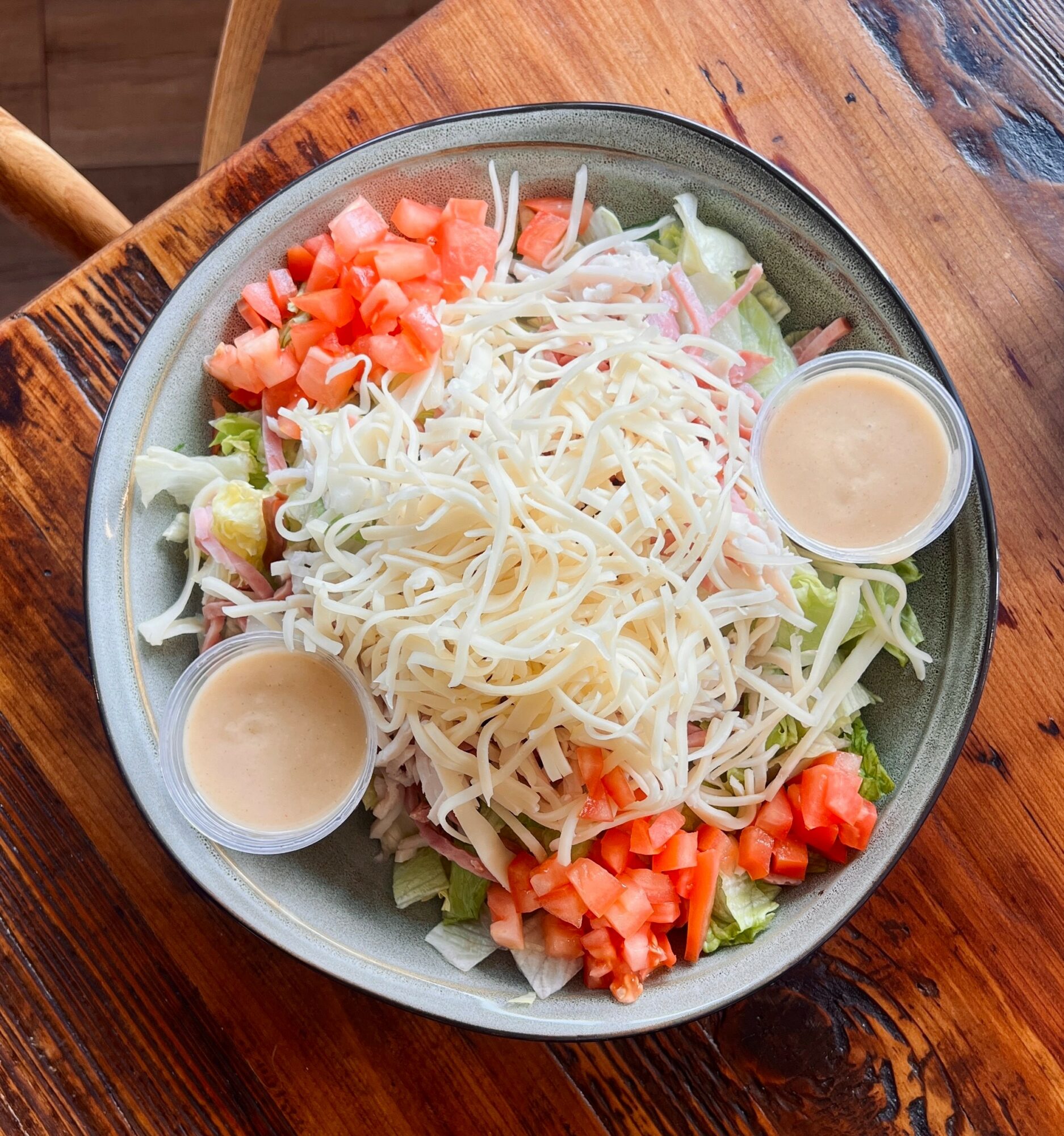 Bowl of salad with shredded cheese, chopped tomatoes, lettuce, and two small cups of dressing on a wooden table.