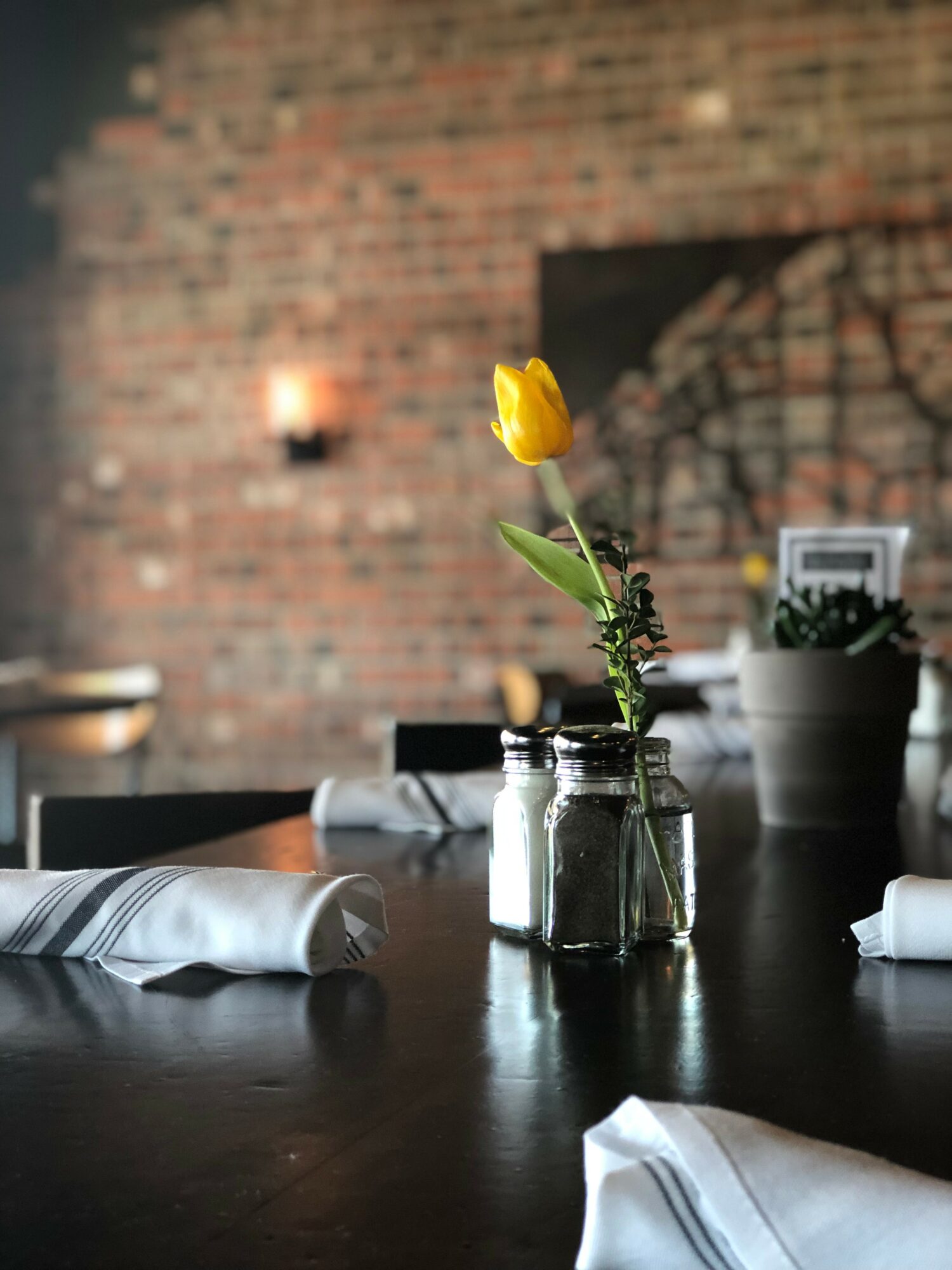 Yellow tulip in a glass jar on a dark table with white napkins, brick wall background, warm lighting.