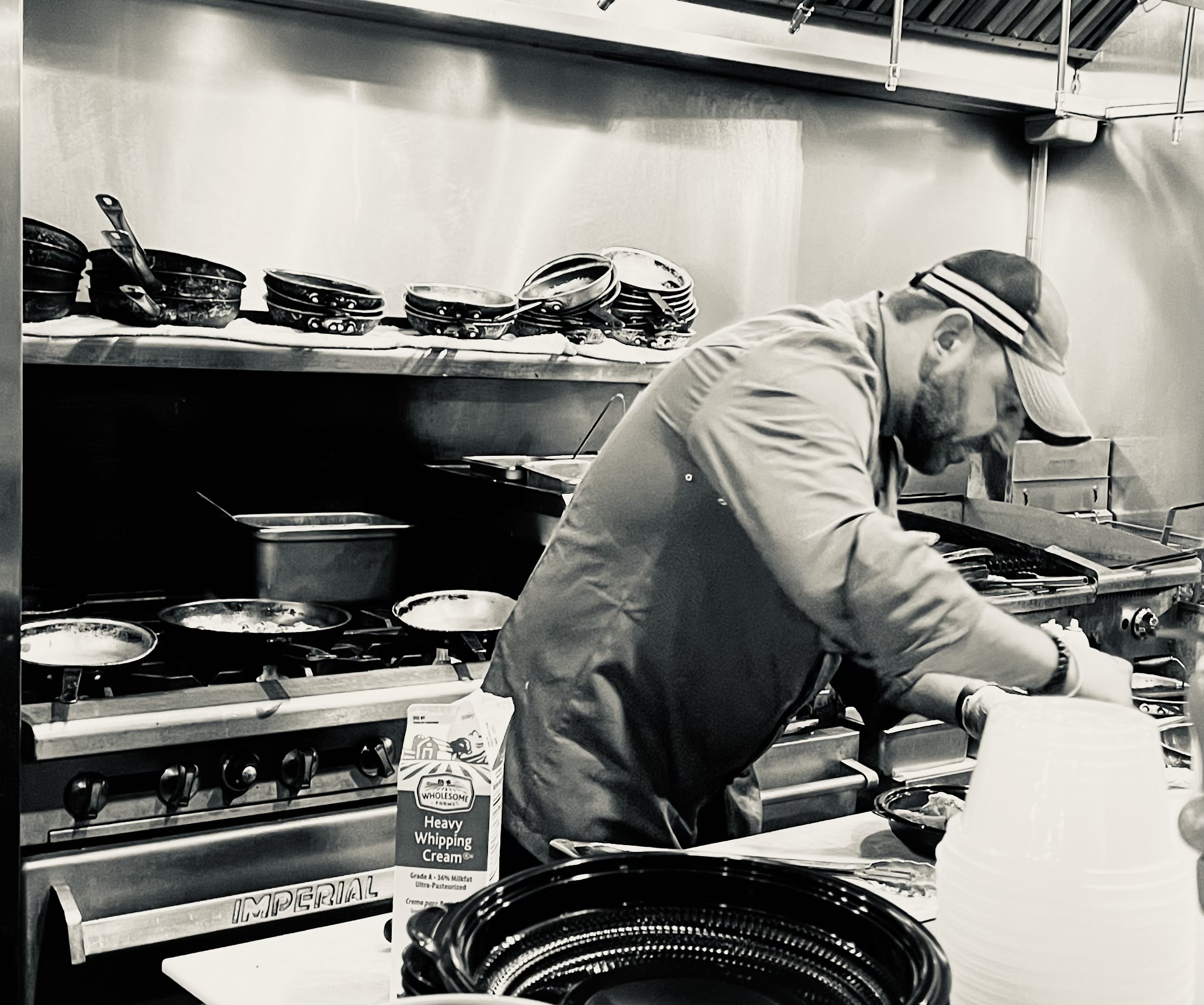 Man cooking in a commercial kitchen, wearing a cap and apron, surrounded by pots, pans, and kitchen utensils.