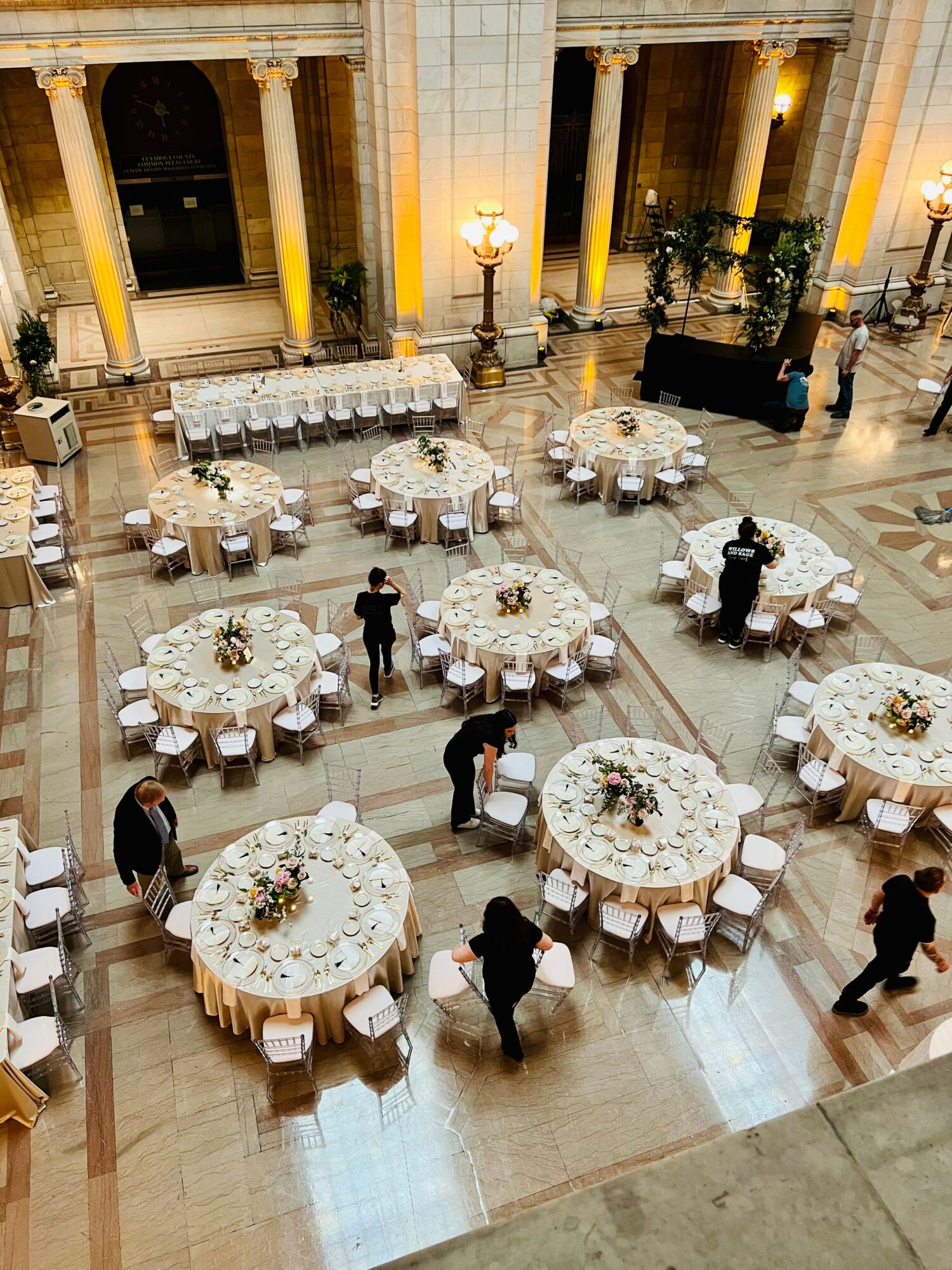 Elegant banquet hall with round tables, floral centerpieces, and people setting up for an event, high ceilings, and tall columns.