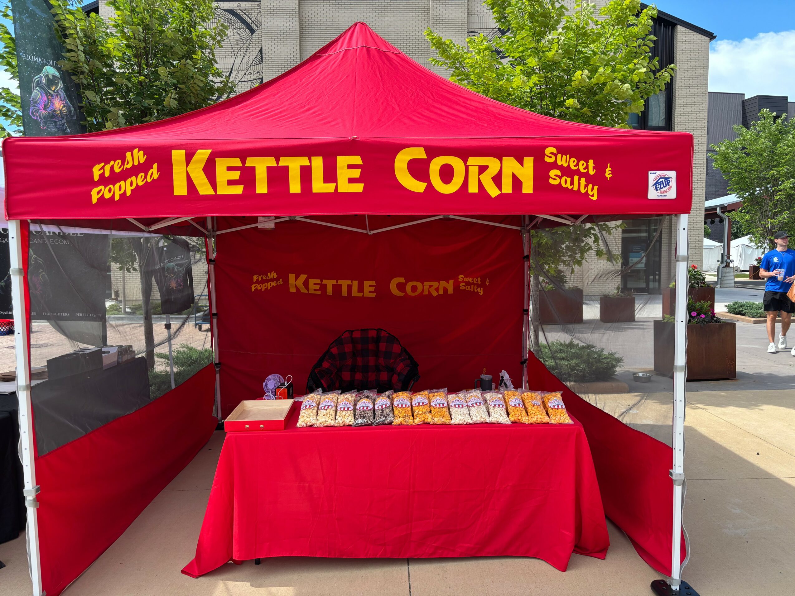 Red tent with yellow and red sign reading Kettle Corn, set up outdoors with a table of popcorn bags.