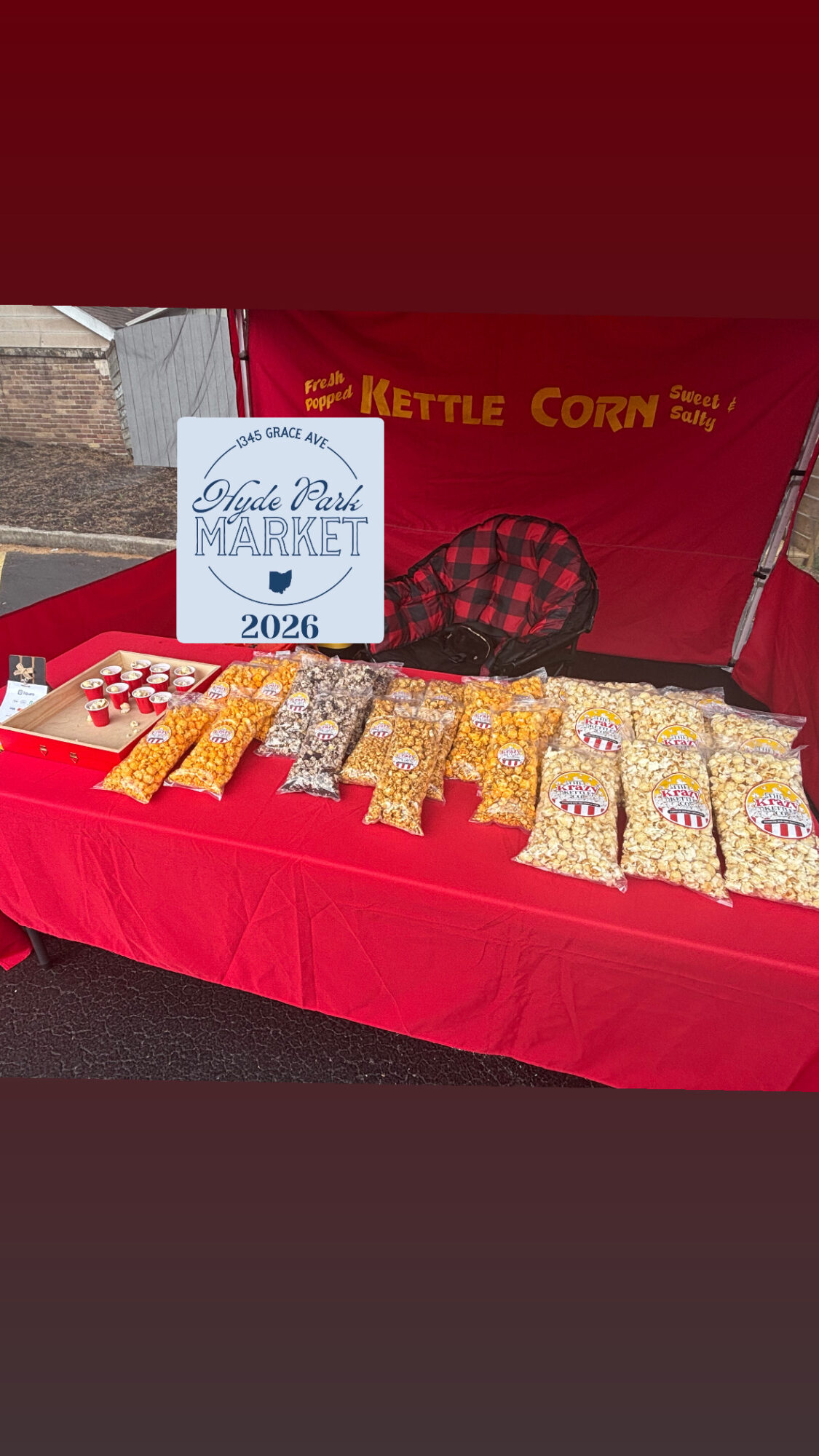 Table with bags of popcorn at a market stall, a sign reading 'Eagle Point Market 2026,' and a red backdrop.