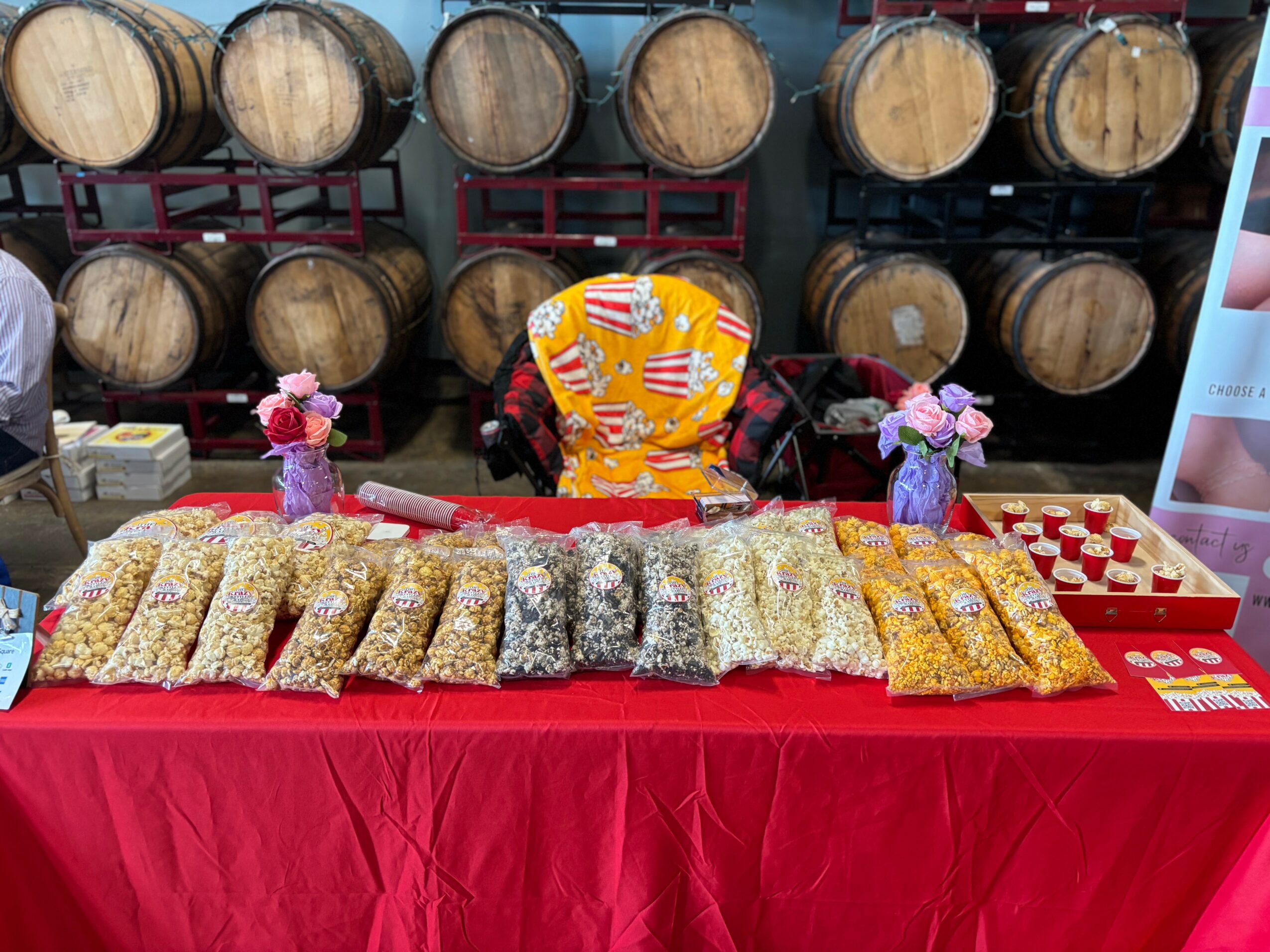 Table with popcorn bags, two small stuffed animals, and a person in a colorful jacket behind barrels, in a store or warehouse.