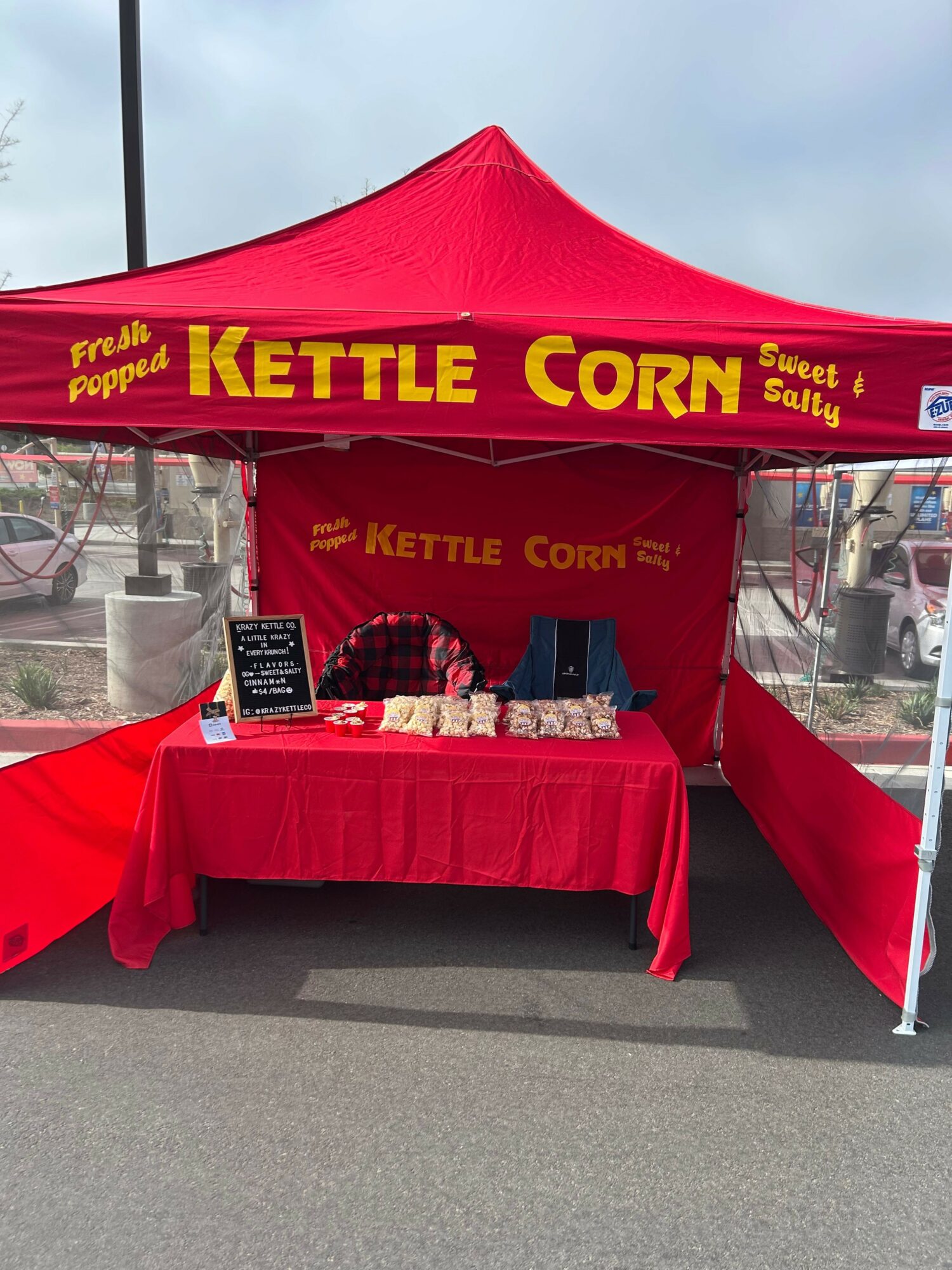 Red tent with yellow text reading 'Kettle Corn' at outdoor market stall, table with popcorn bags, person behind table, overcast sky