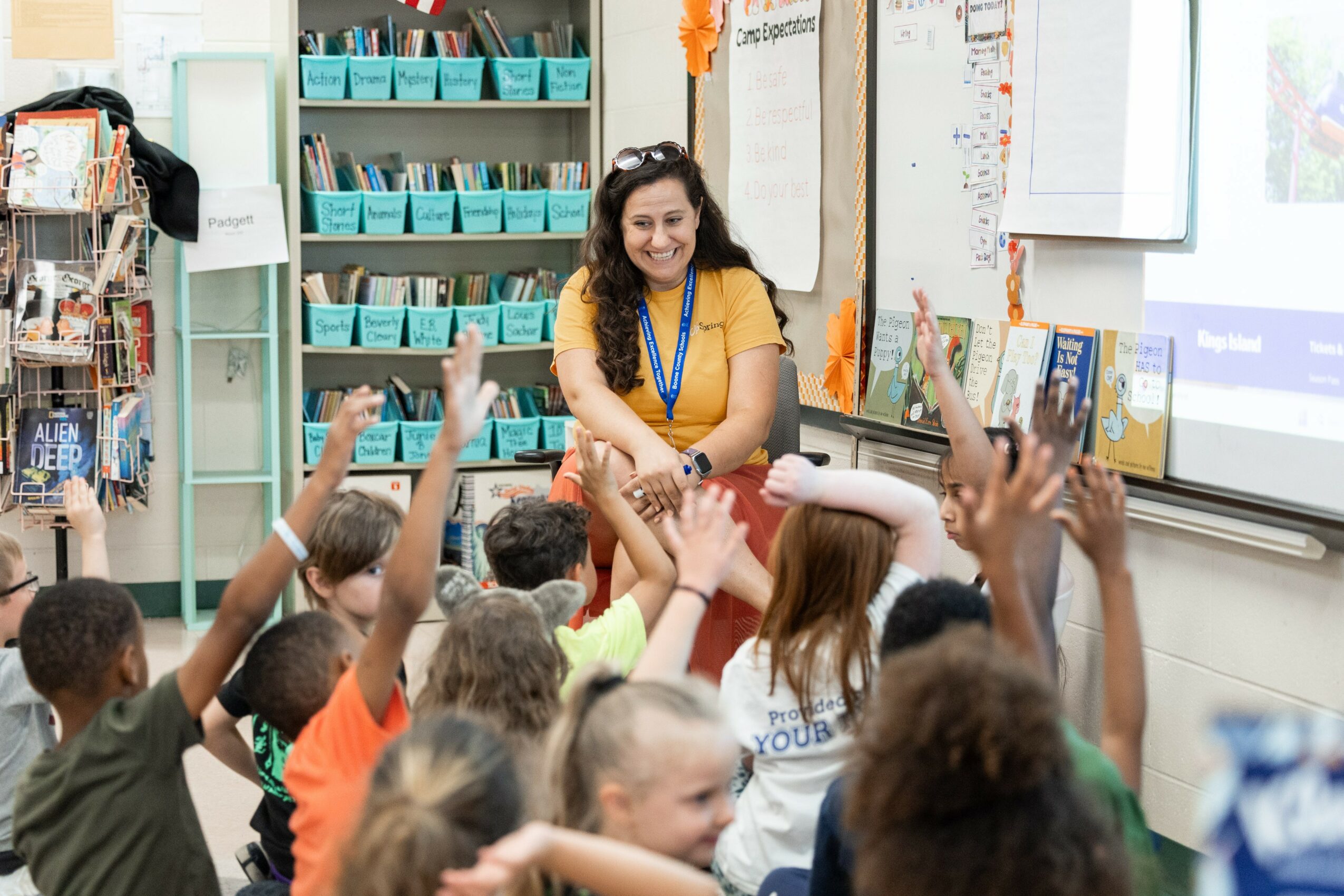 Teacher smiling at students raising hands in classroom, educational materials on walls and shelves, students engaged and attentive.