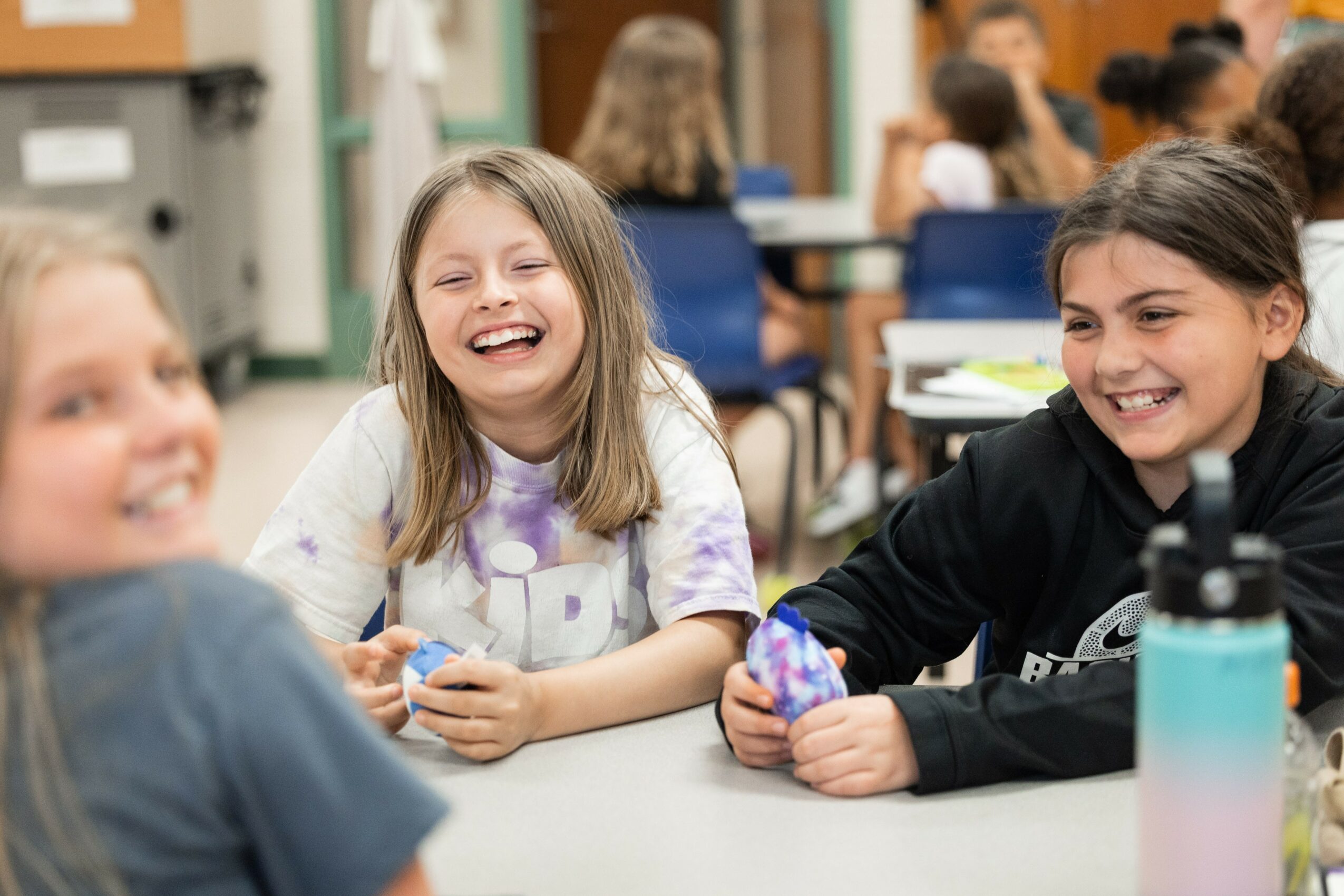 Three children laughing and smiling at a table in a classroom, with other students in the background.