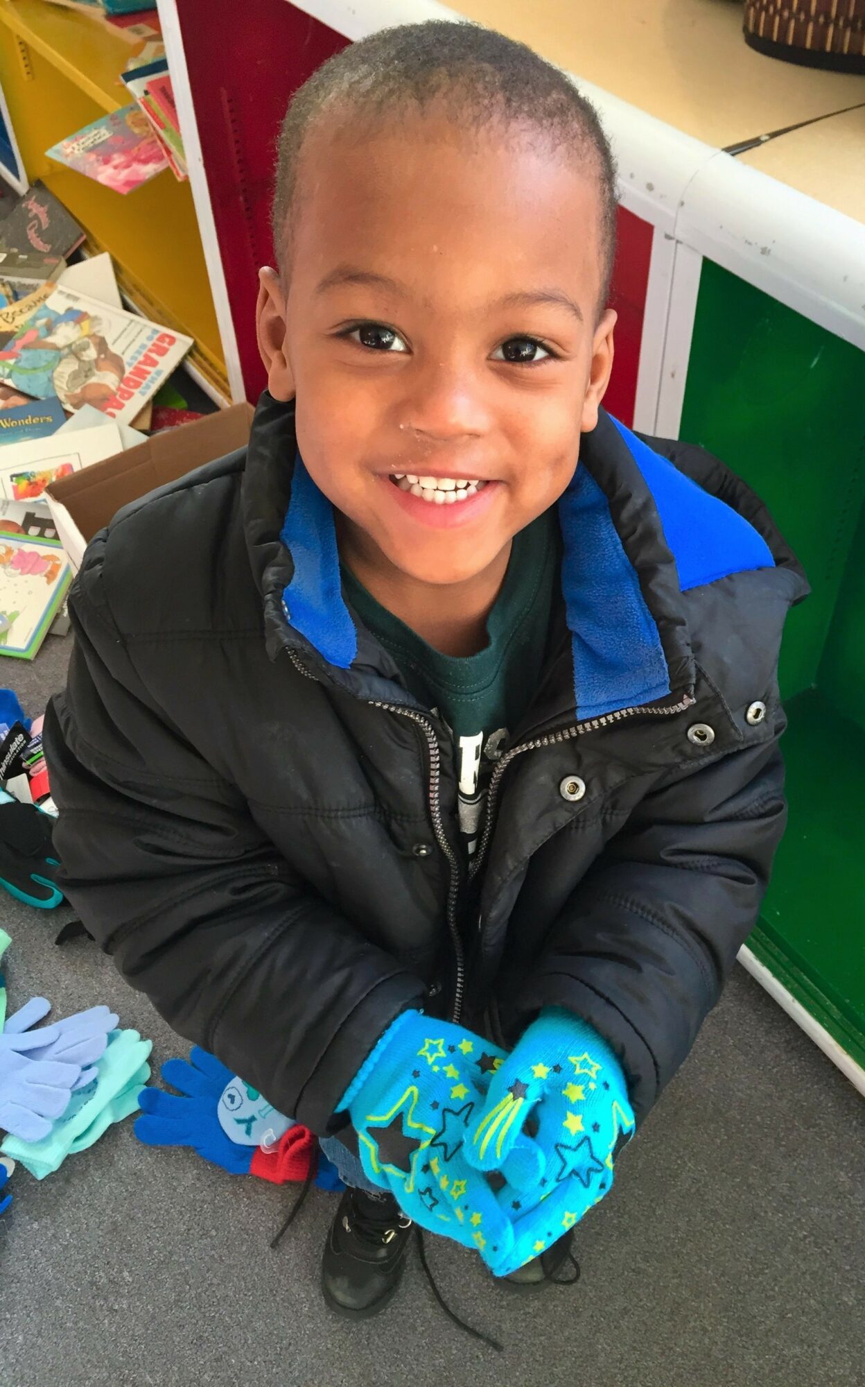 Young boy with short hair smiling, wearing a black jacket and blue gloves, in a colorful indoor setting.