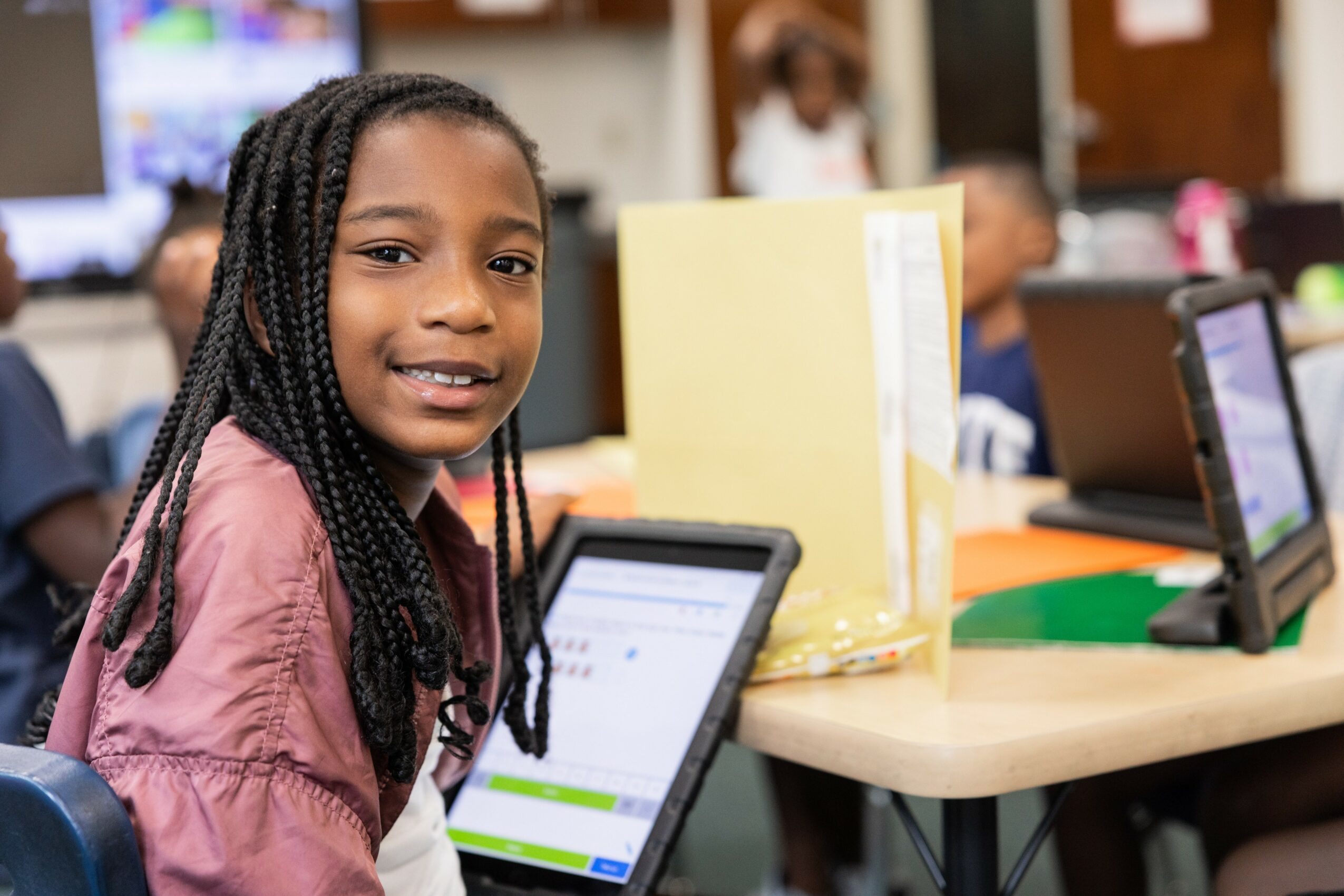 Young girl with braided hair smiling in classroom with tablets and books, background shows other students and classroom setting.