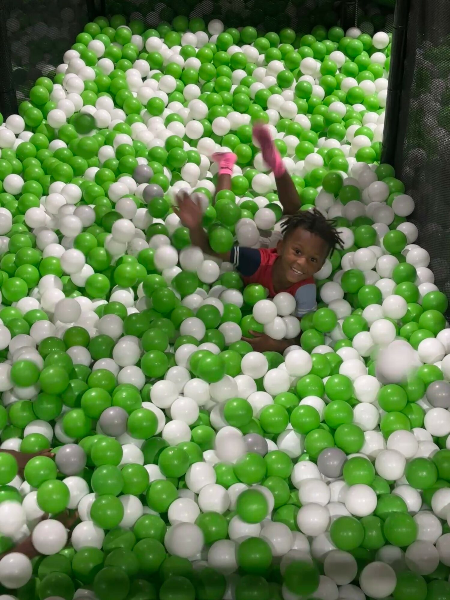 Child playing in a ball pit filled with green, white, and a few pink balls, smiling and raising arms.