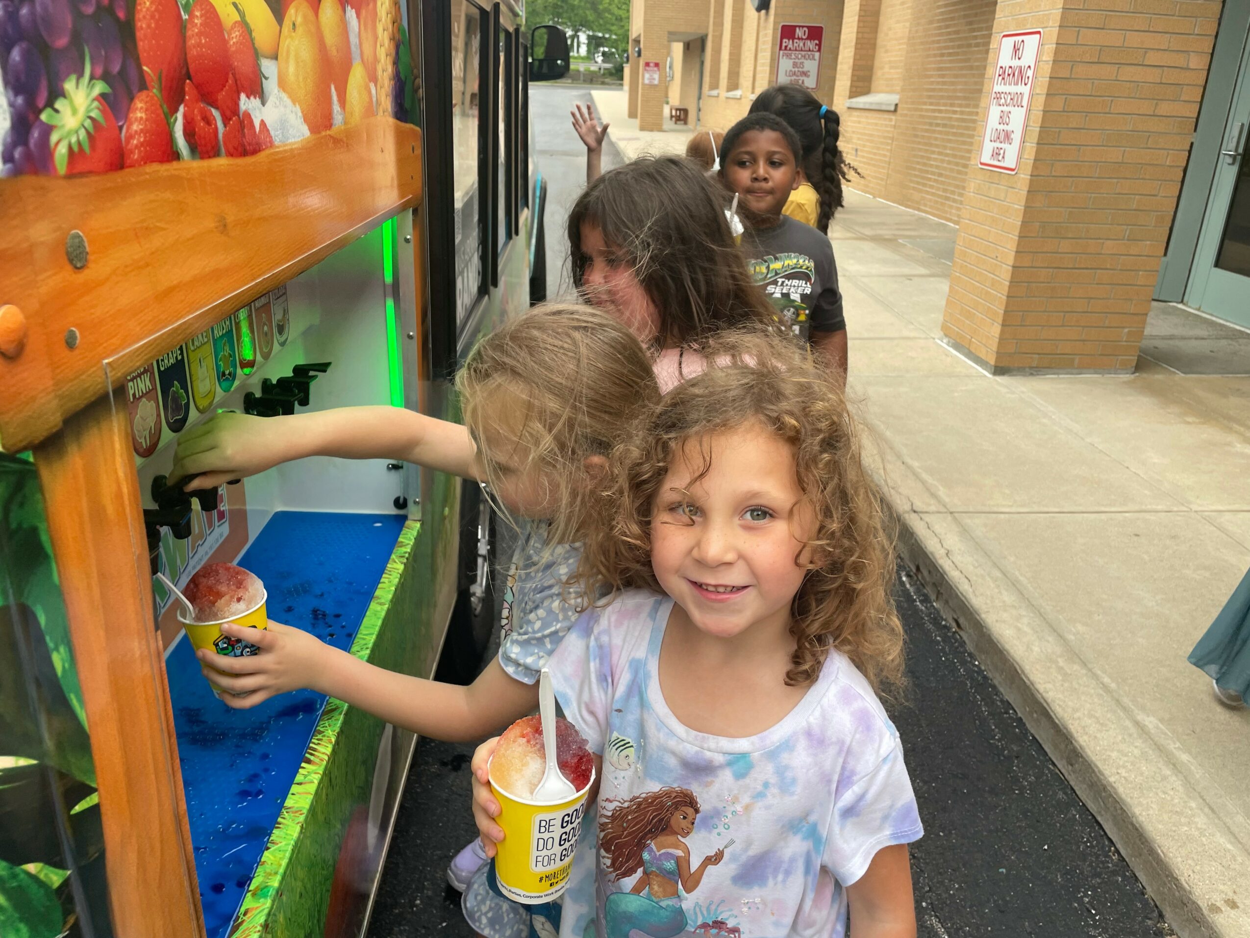 Group of children at a vending machine outside, one girl smiling and holding a snack, others selecting items.