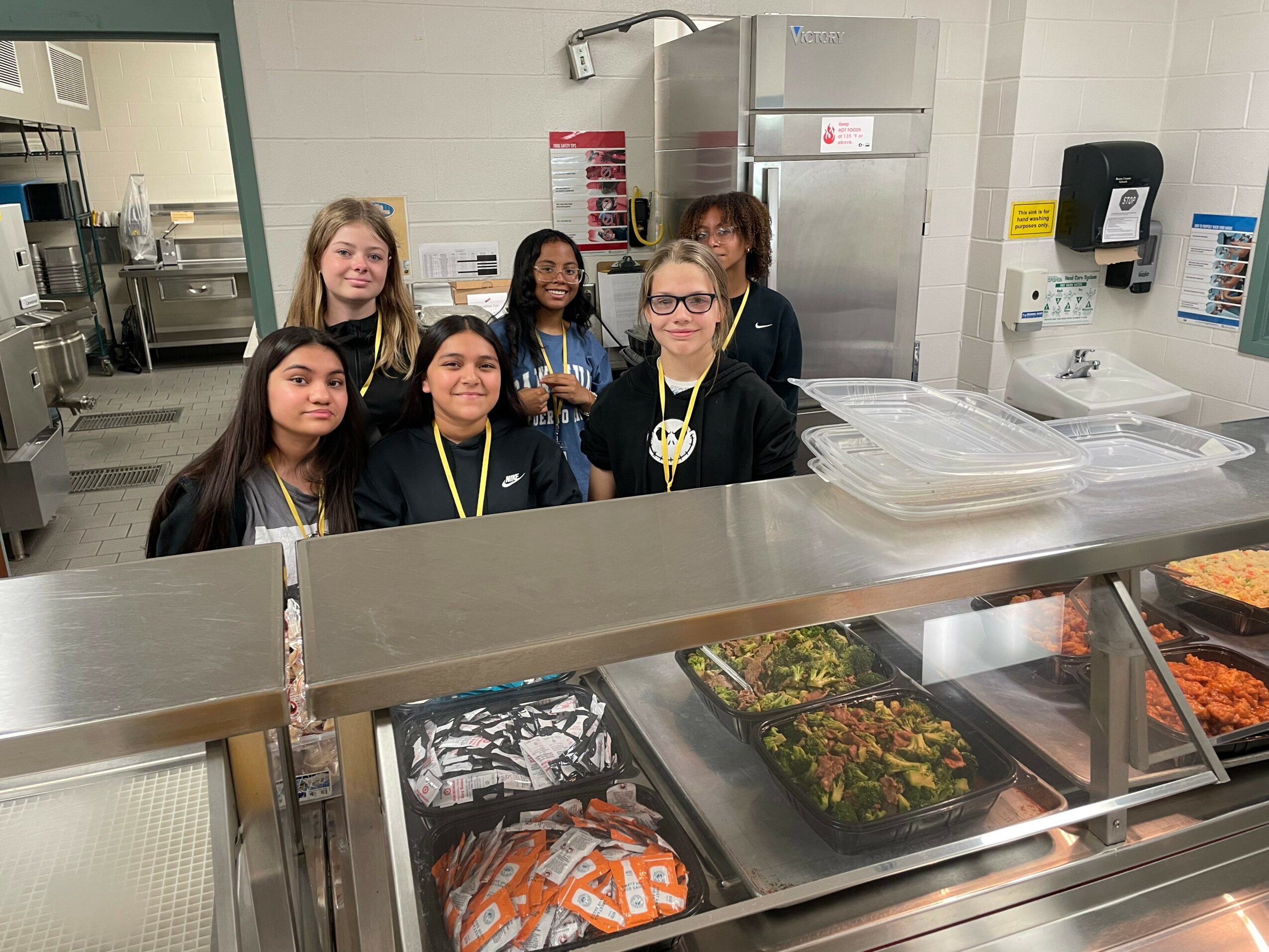 Six children standing behind a cafeteria serving counter with food trays, smiling in a kitchen setting.