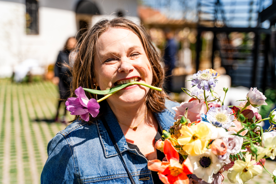 Woman smiling with a flower in her mouth, holding a bouquet of colorful flowers outdoors.