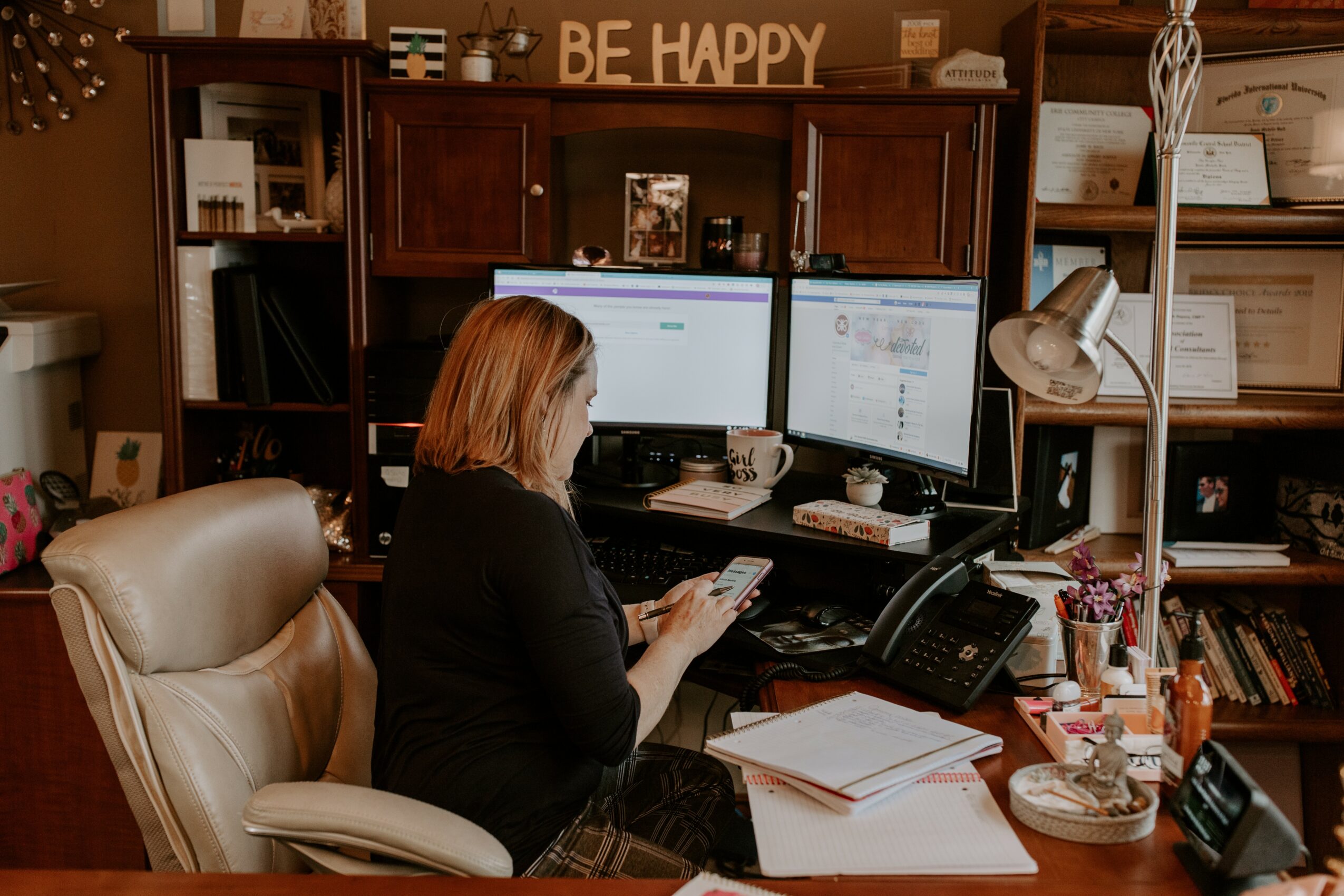 Woman sitting at desk with two computer monitors, using a smartphone, surrounded by papers and office supplies, bookshelf behind.