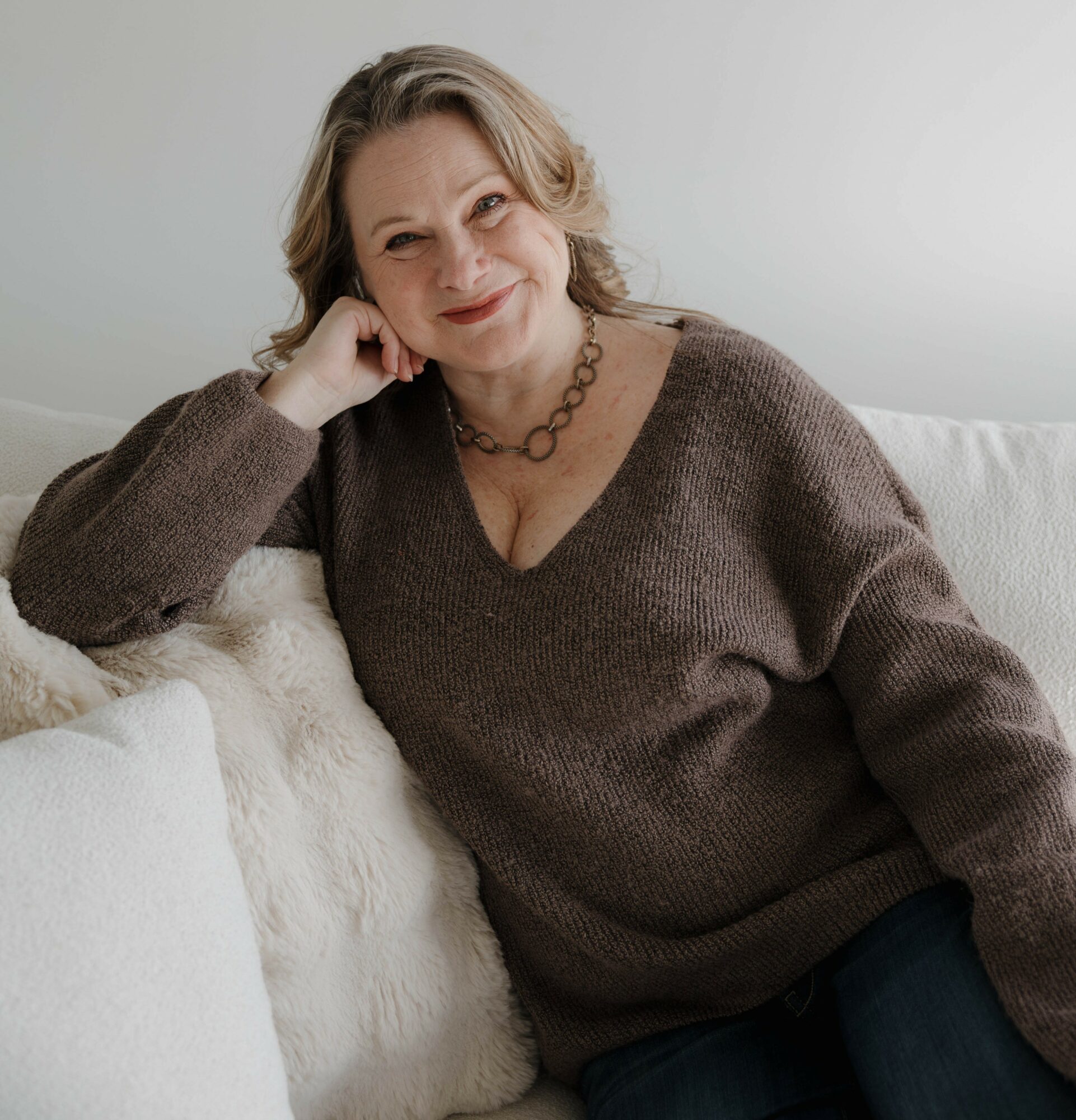 A woman with shoulder-length gray hair, wearing a brown sweater and necklace, sitting on a white couch, smiling gently.
