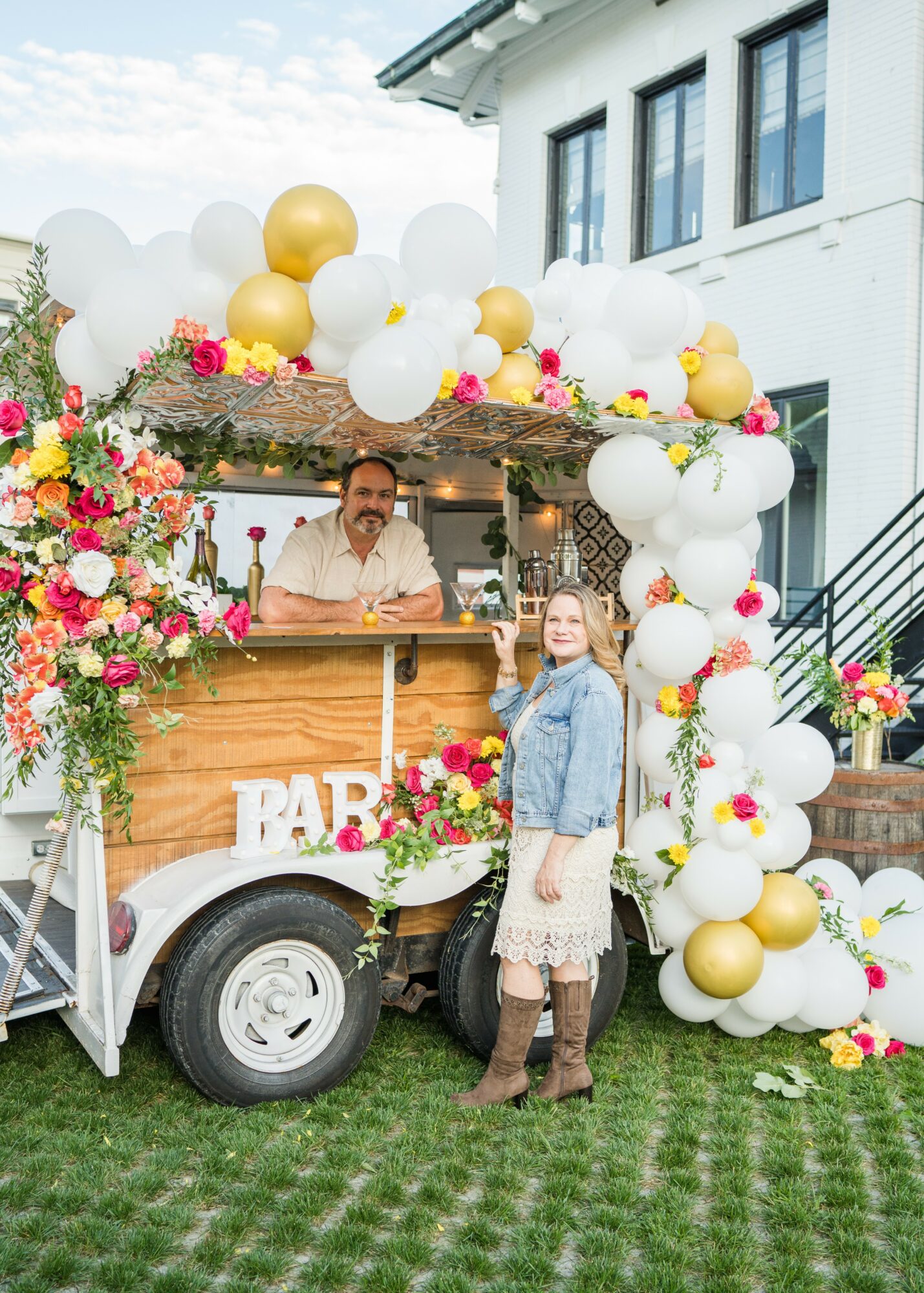 A woman stands next to a decorated bar cart with balloons and flowers, smiling outdoors in front of a white building.