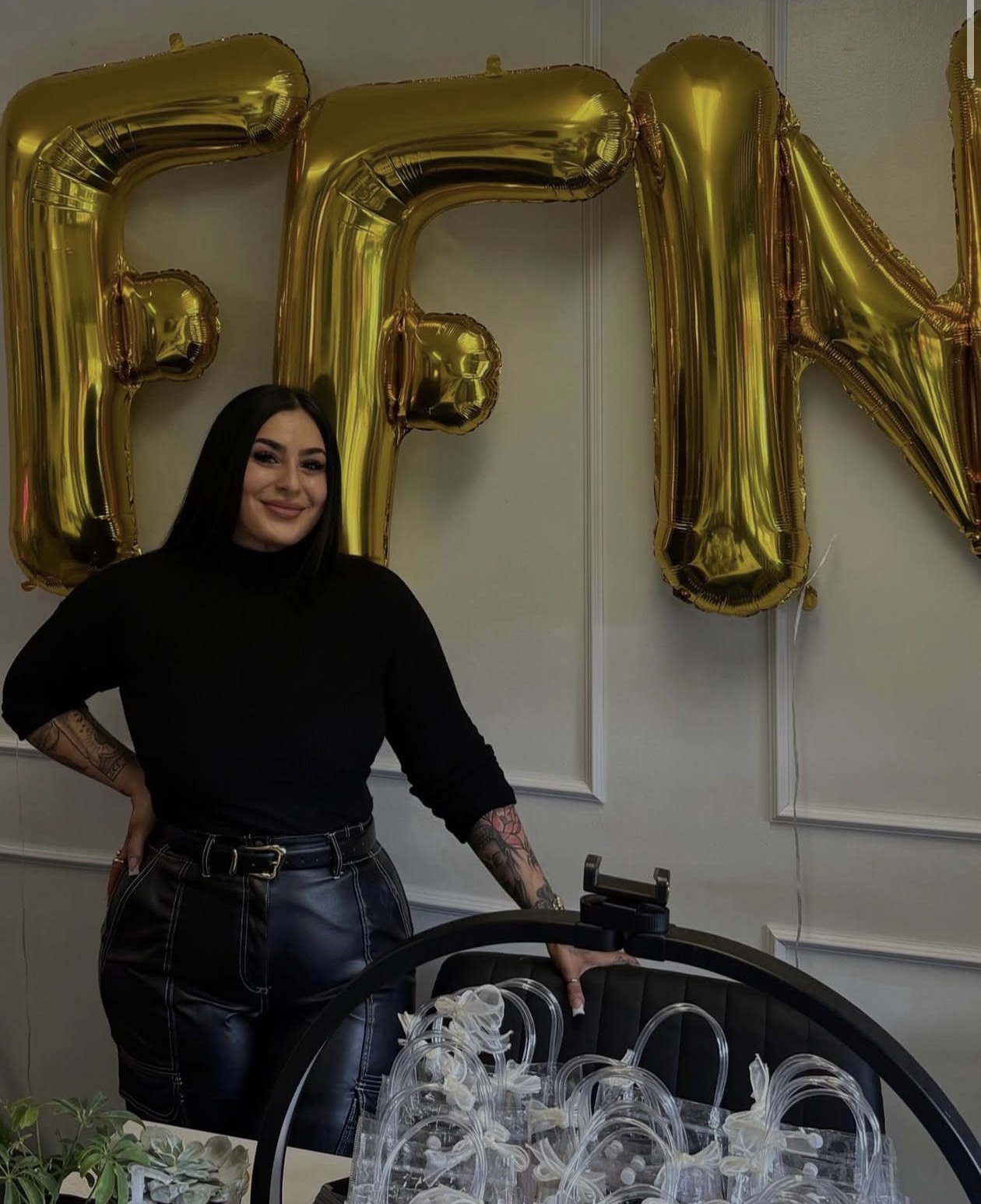 Woman with dark hair and black outfit standing in front of gold balloon letters, smiling, with a white wall background.