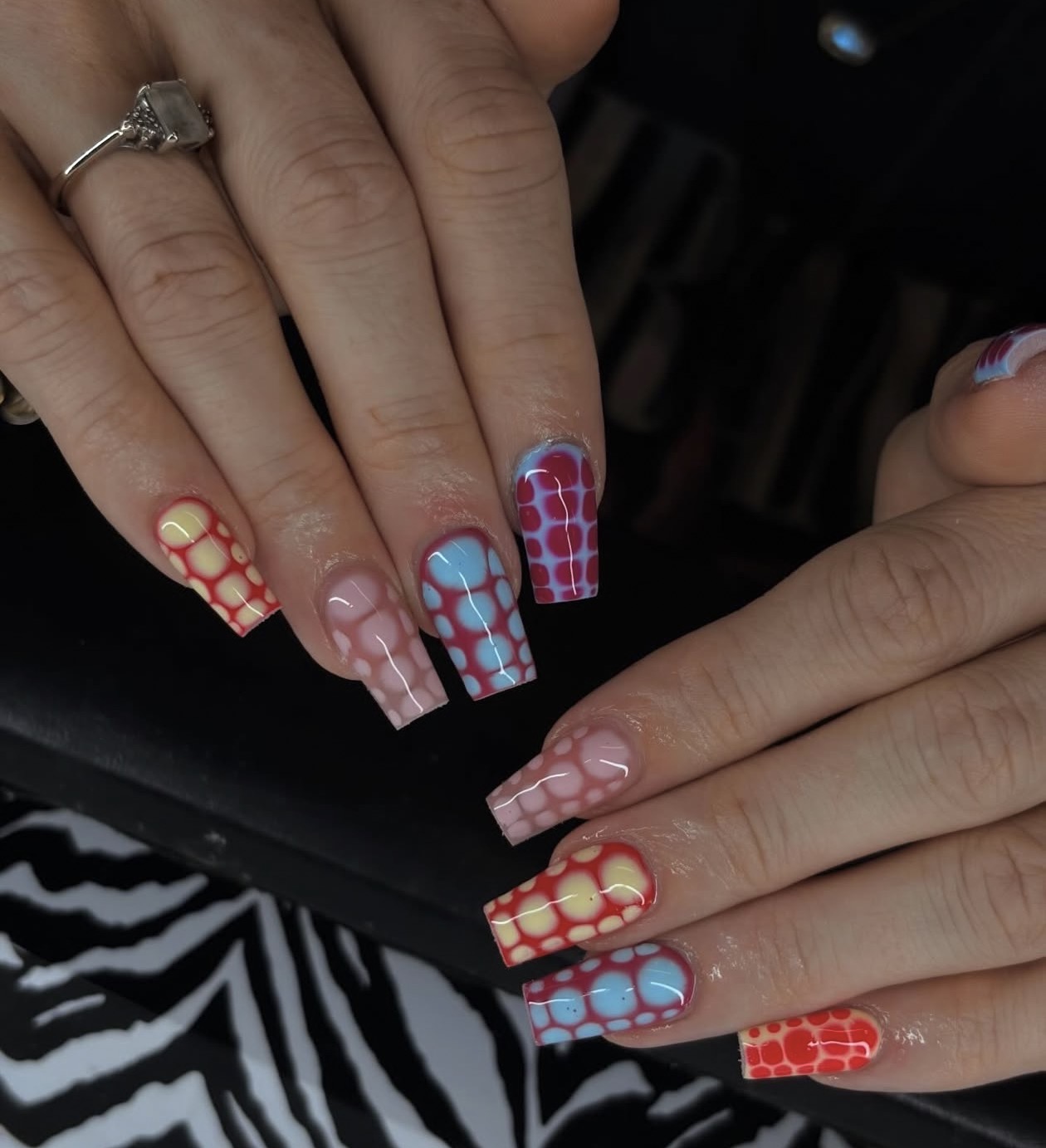 Hands with colorful patterned nail art, rings, and a black and white patterned fabric background.