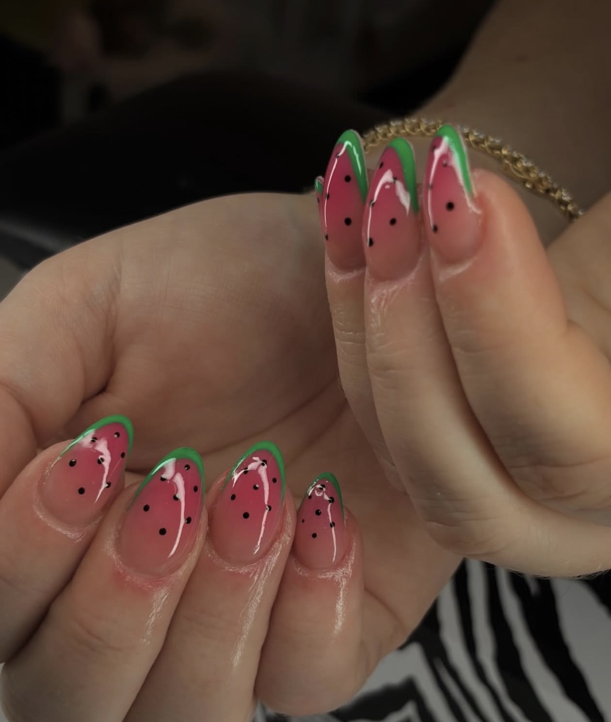 Close-up of hands with watermelon-themed nail art, pink nails with black seeds and green rind design, gold bracelet visible.