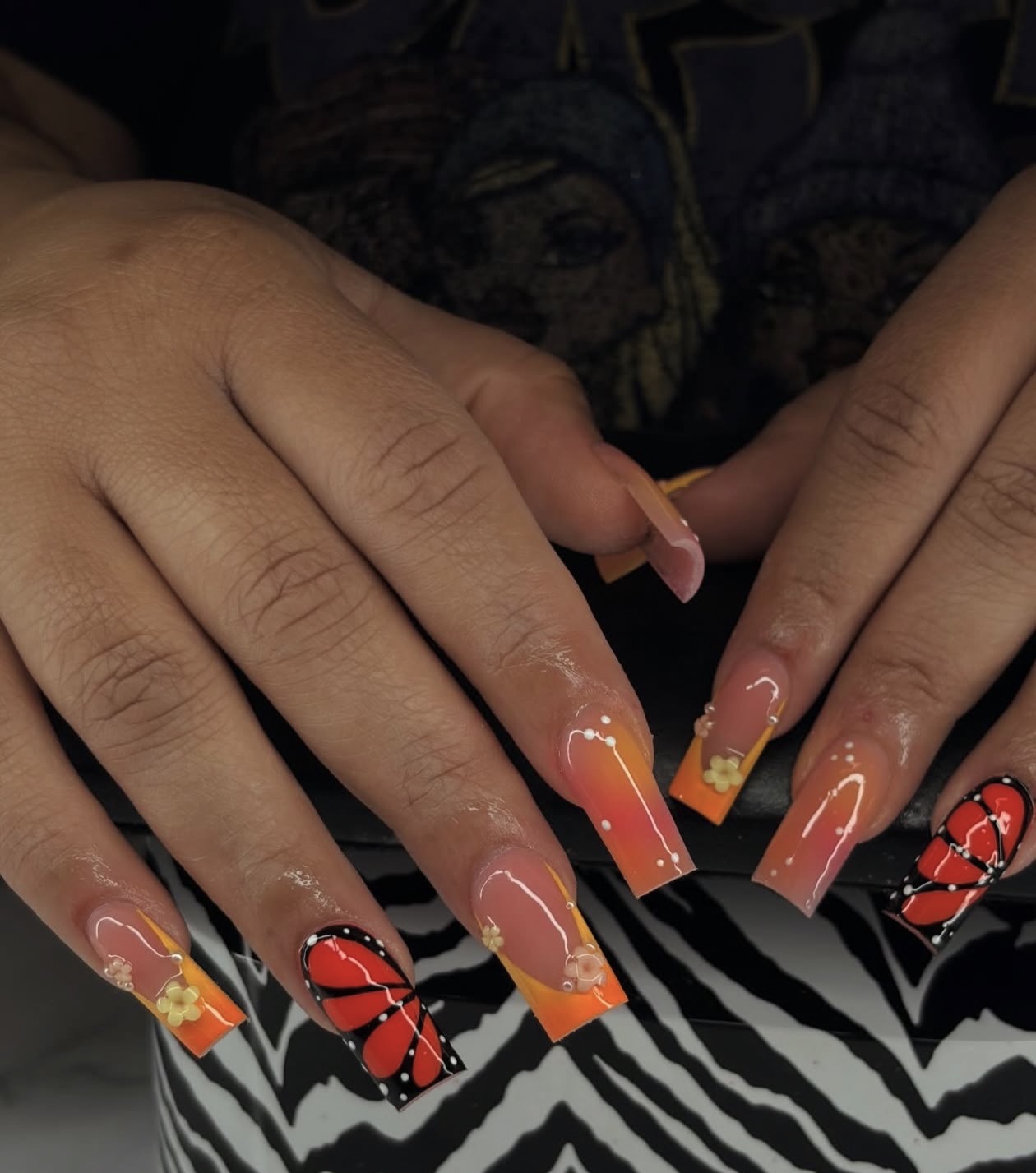 Hands with decorated nails, featuring butterfly designs and orange tips, resting on a black and white striped surface.
