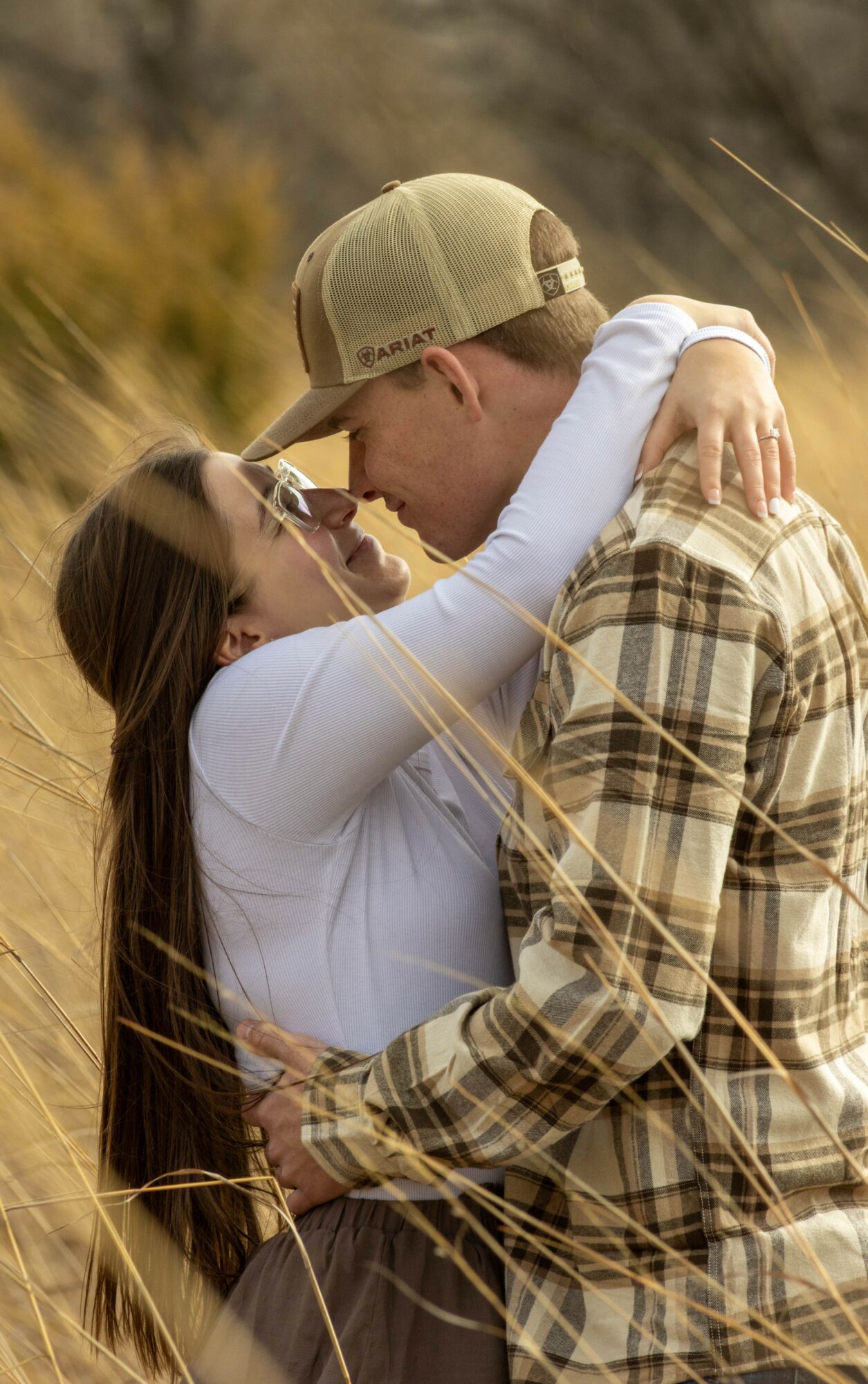A couple embraces outdoors, with the woman holding the man and looking at him, in a natural setting with tall grass.