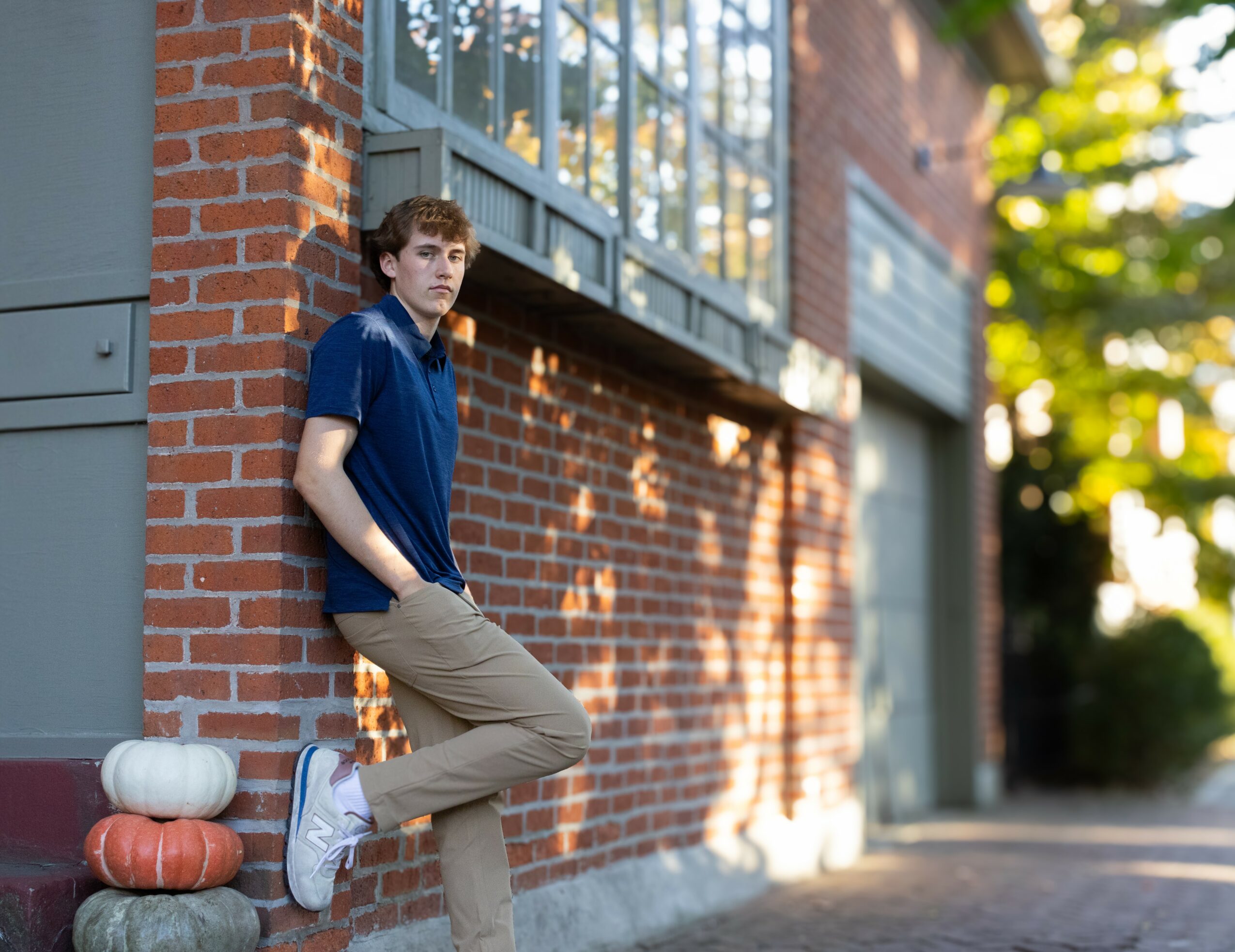 Young man leaning against a brick wall with stacked pumpkins nearby, outdoors in sunlight.