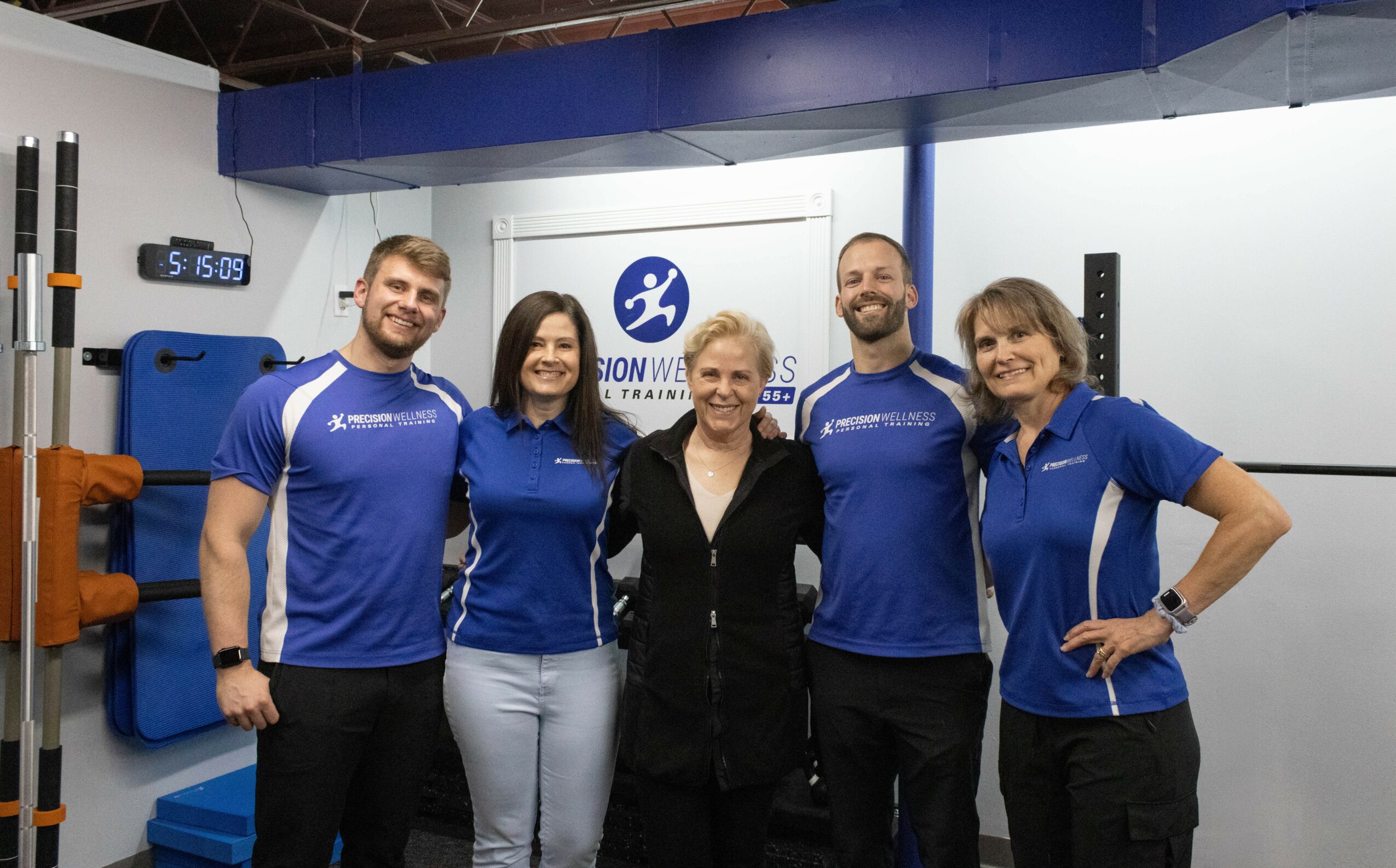 Group of five people standing indoors, smiling, with a logo and equipment in the background.