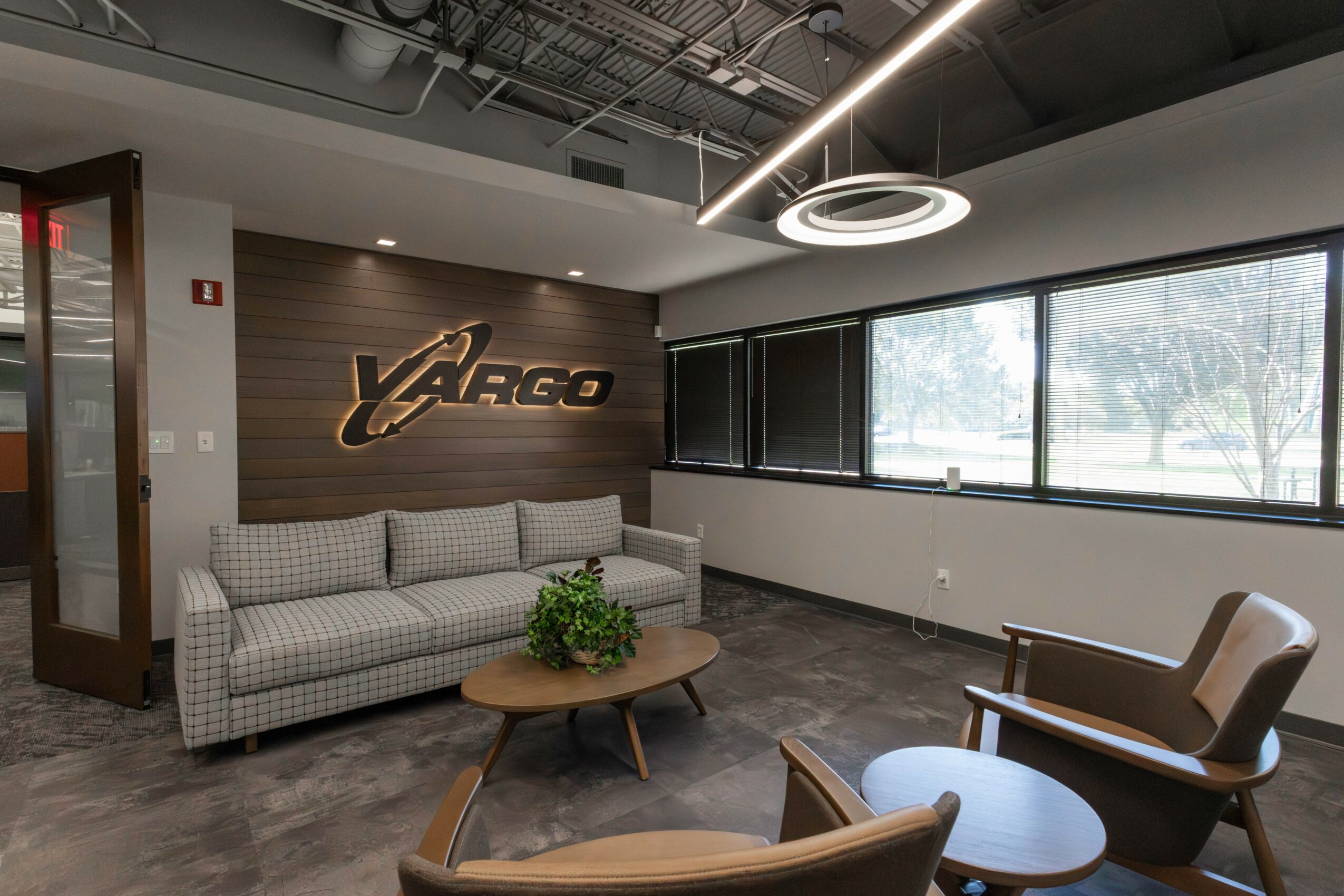 Modern office lounge with a gray sofa, wooden coffee table with plant, and a brown armchair near large windows.