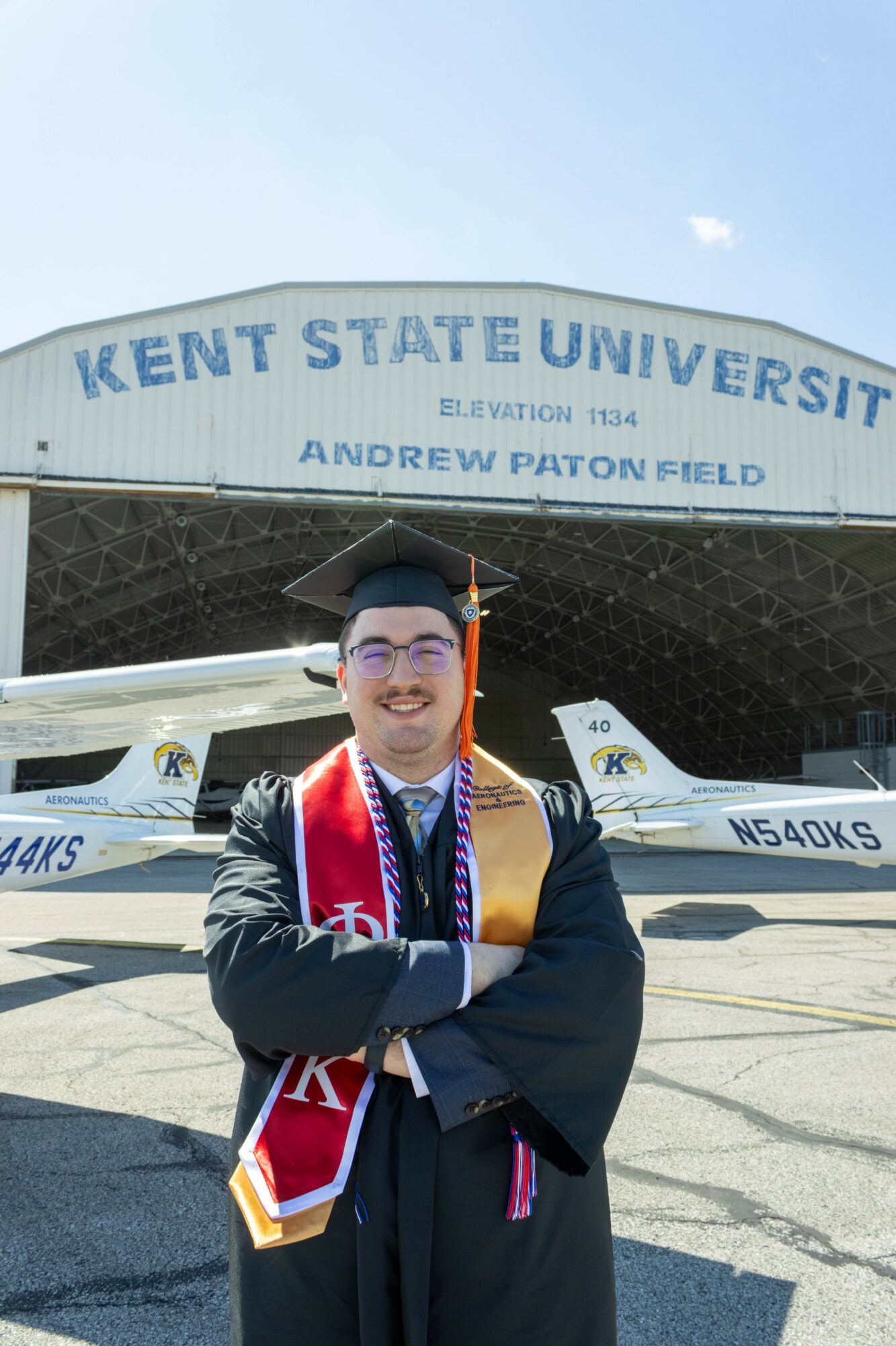 Graduate in cap and gown smiling in front of Kent State University sign and aircraft.