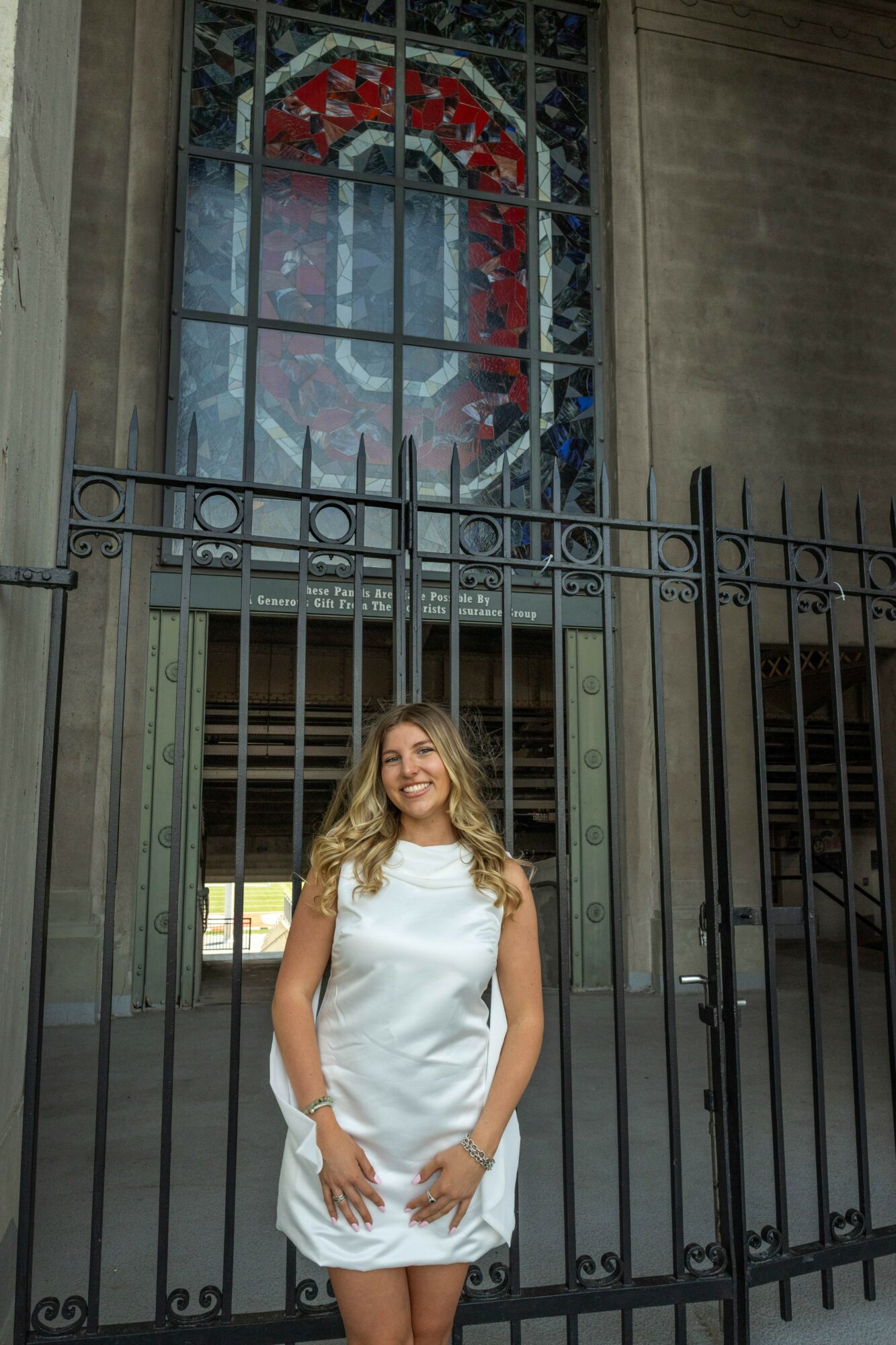 Woman in white dress standing behind a black metal fence in front of a stained glass window with a red and green design.
