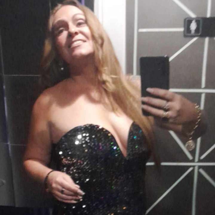 Woman with long hair taking a mirror selfie, wearing a strapless, sequined dress, smiling, in a modern room.