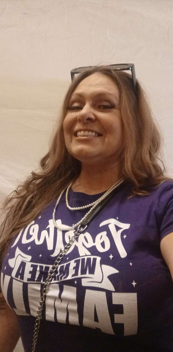 Woman smiling, wearing glasses on head, purple T-shirt with white text, and a necklace.