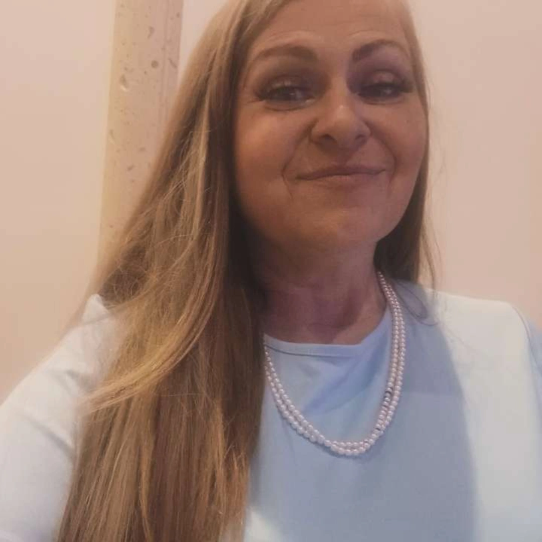 Woman with long hair smiling, wearing a light blue top and pearl necklace, indoors.