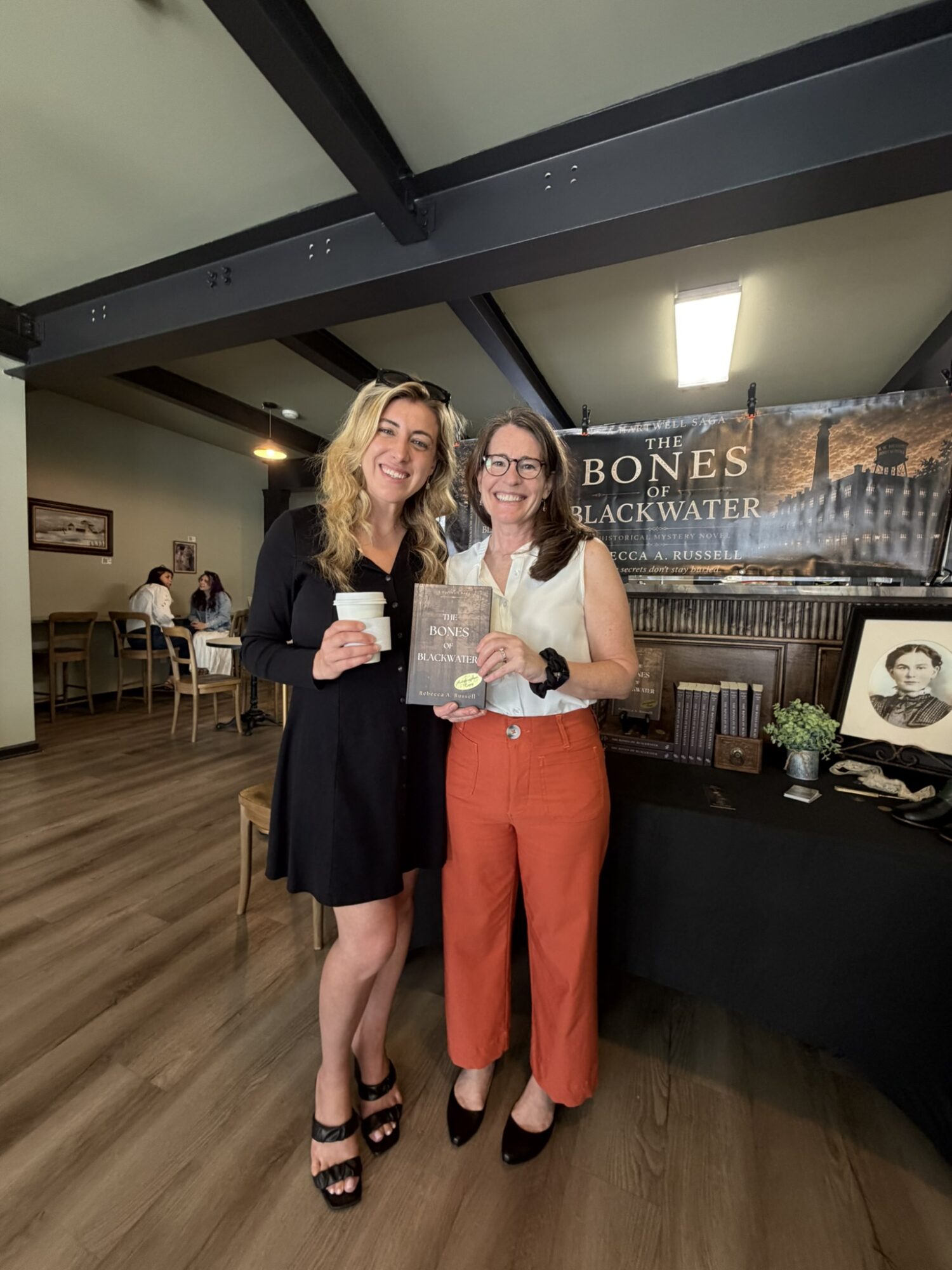 Two women standing indoors, smiling, holding a book and a cup, with a banner and table in the background.