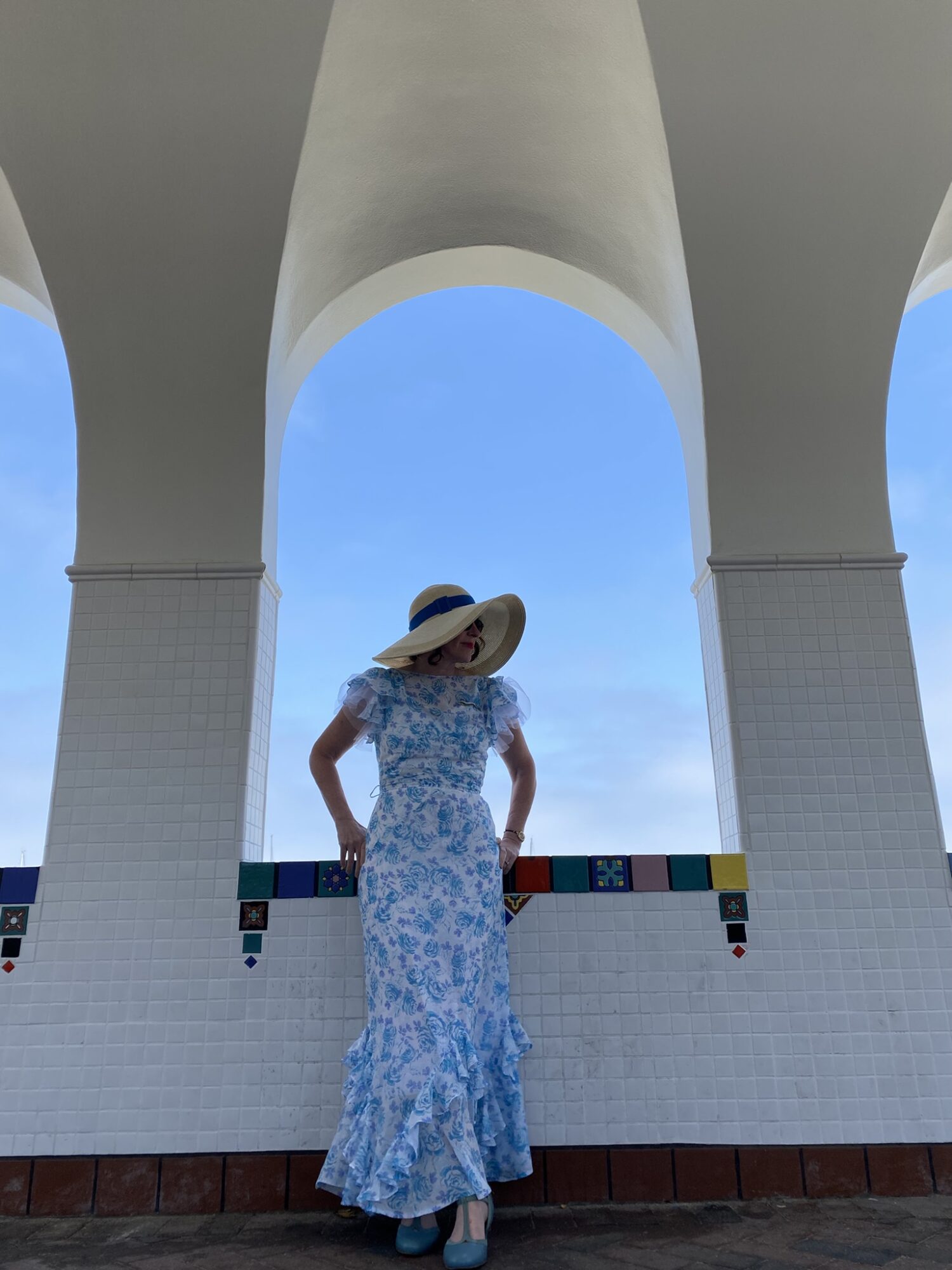 Woman in a floral dress and large hat standing between white arches with blue sky background.