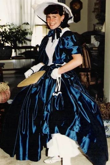 Young girl in a blue Victorian-style dress with white gloves and parasol, smiling indoors with plants and furniture nearby.