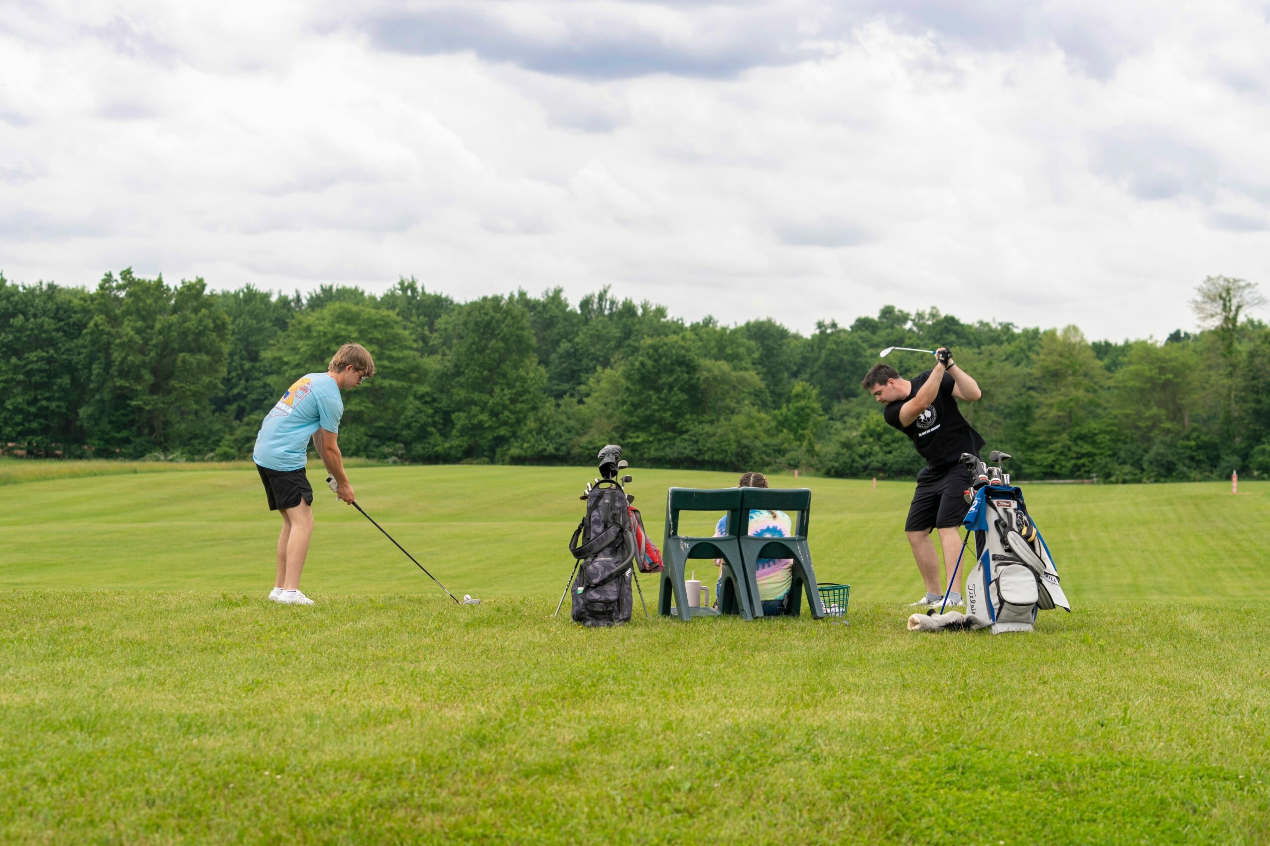 Two people on a golf course, one preparing to swing and the other setting up a shot, with golf bags and a small table nearby.