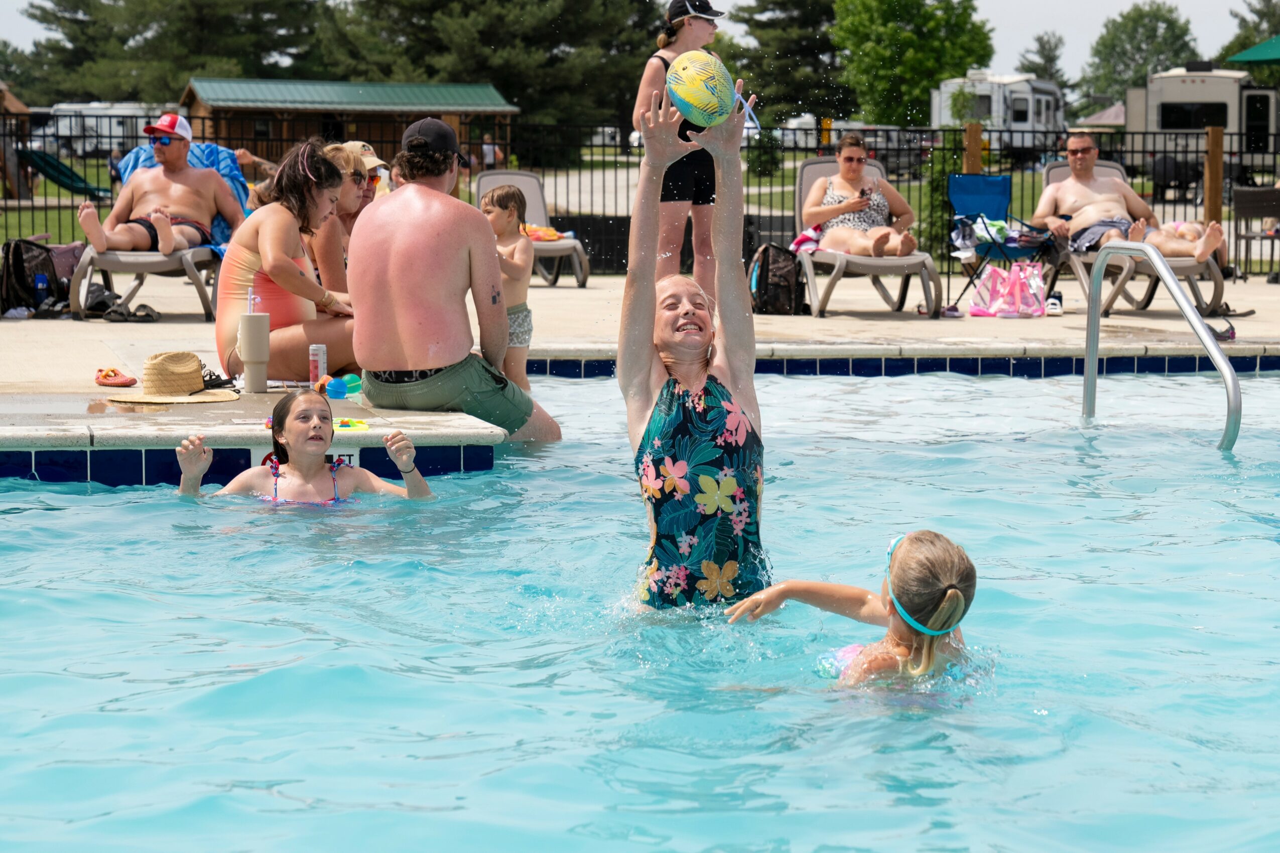 Children and adults in a swimming pool with lounge chairs and trees in background, some playing with a ball, sunny day.