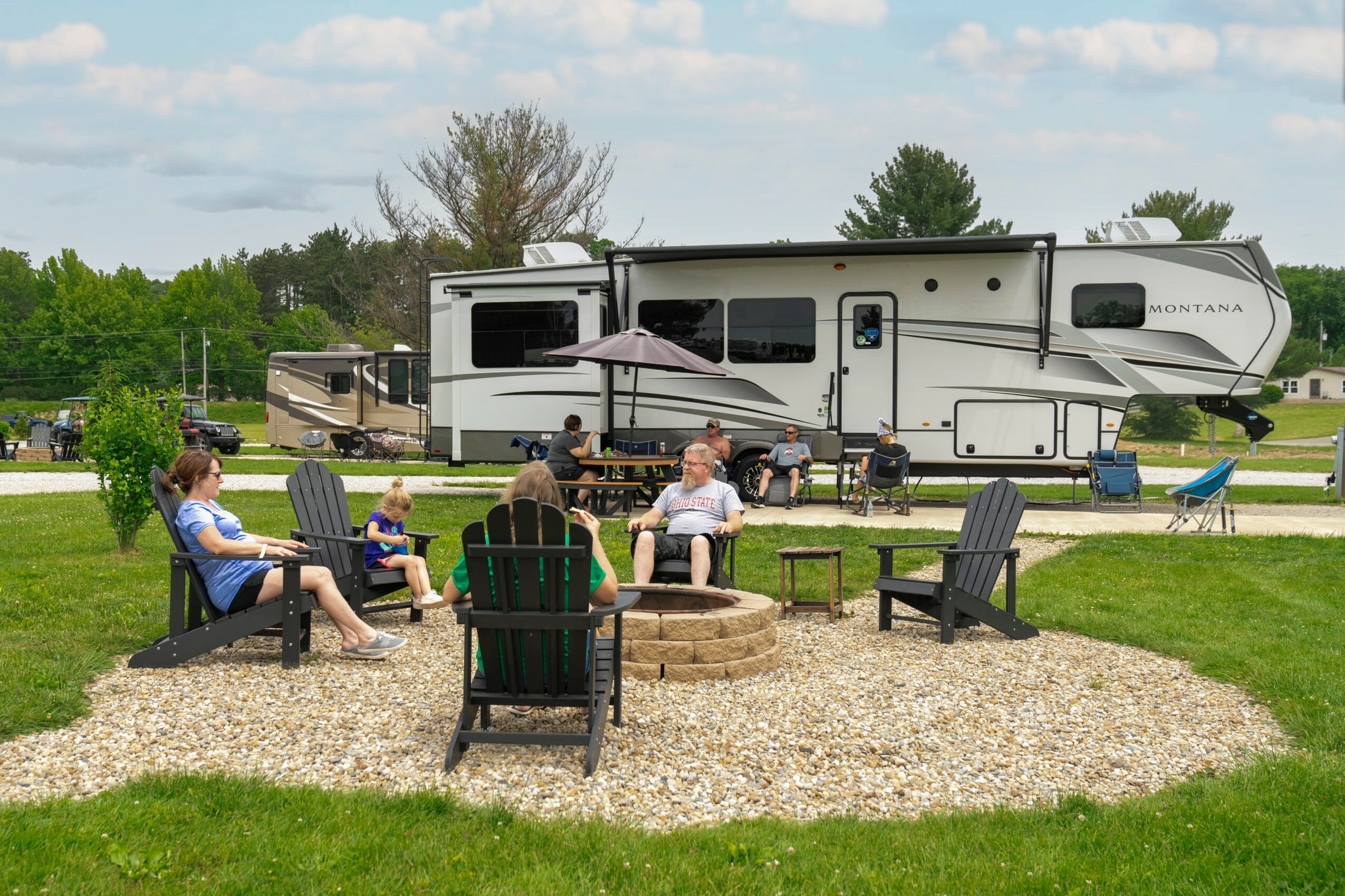 Group of people sitting around a fire pit with chairs on a gravel area, with an RV in the background, outdoors.